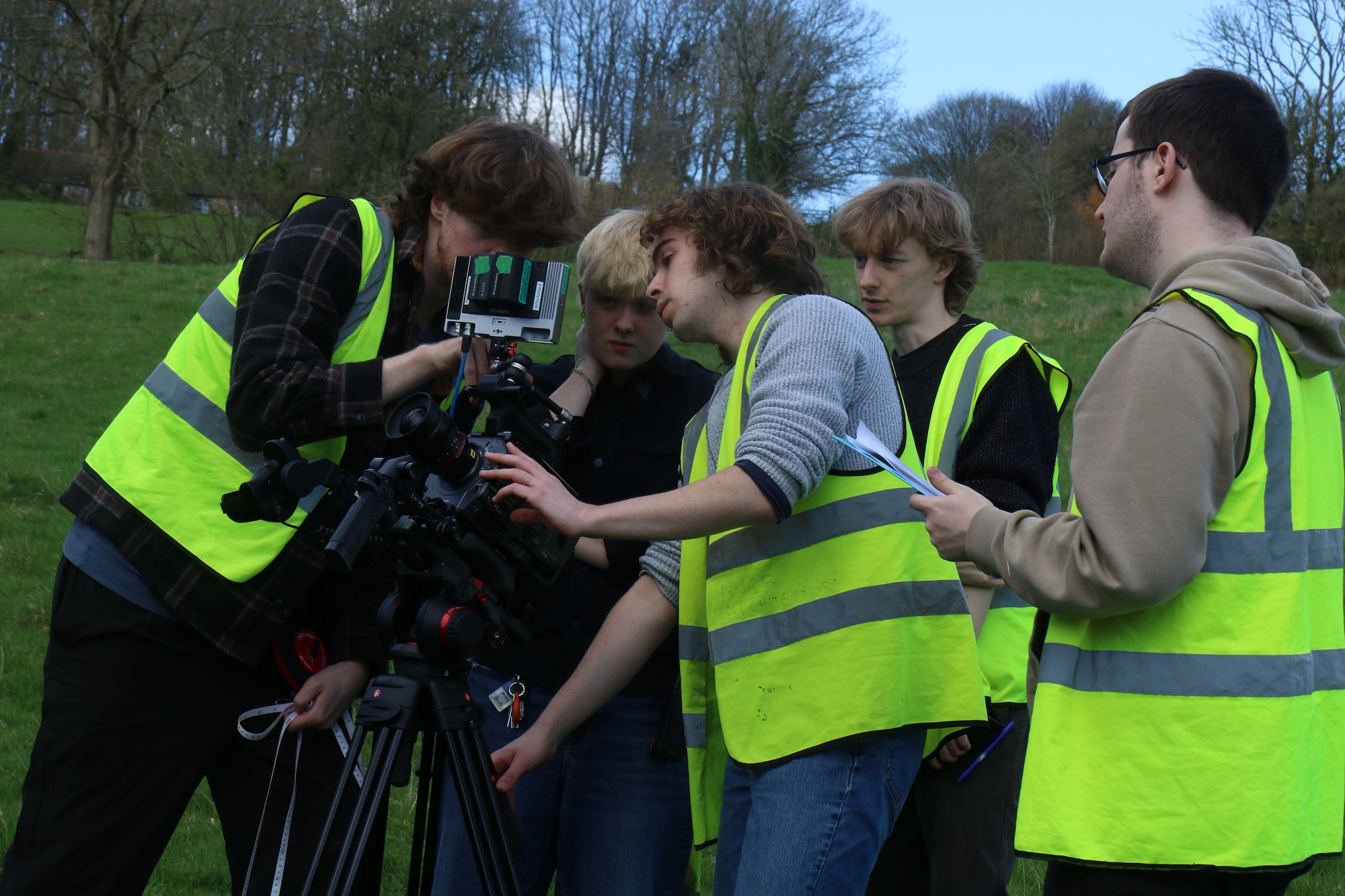 A group of five individuals wearing yellow high-visibility vests are gathered around a video camera mounted on a tripod. One person is adjusting the camera while others observe, with greenery visible in the background.