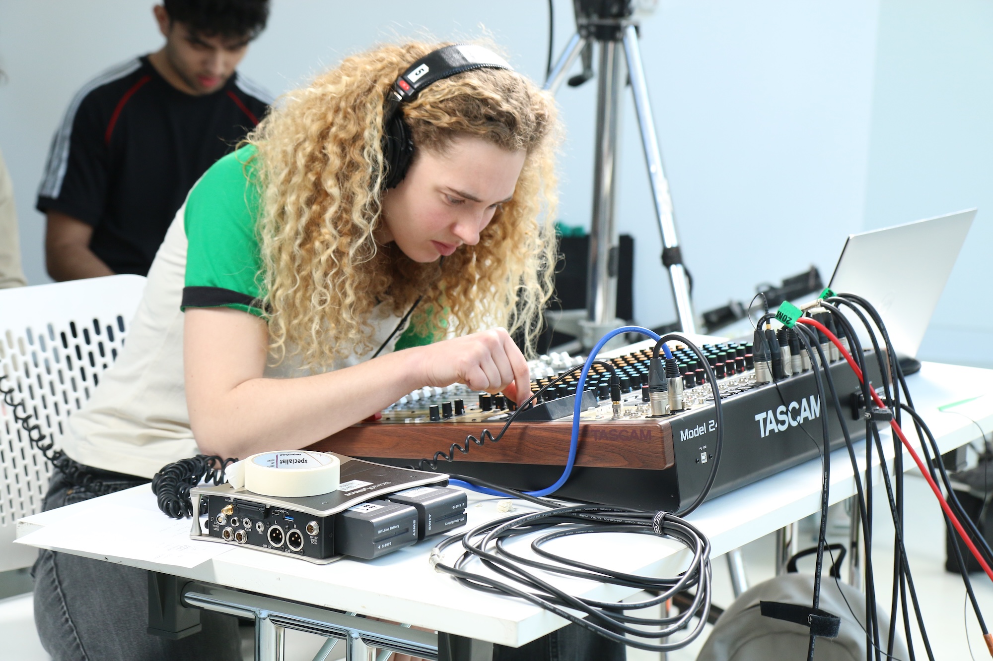 An Arts University Bournemouth student is focused on adjusting a mixing console with various knobs and cables. In the background, another individual is sitting at a table, and there are studio equipment and a laptop visible nearby.