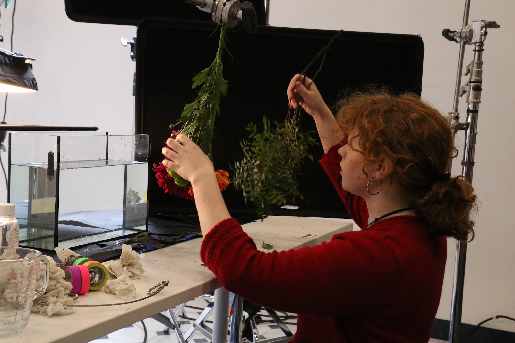 A woman with curly ginger hair holds two leafy green branches above a worktable. In front of her, a glass tank filled with water is visible. The workspace contains tools and materials, with soft lighting from overhead lamps.