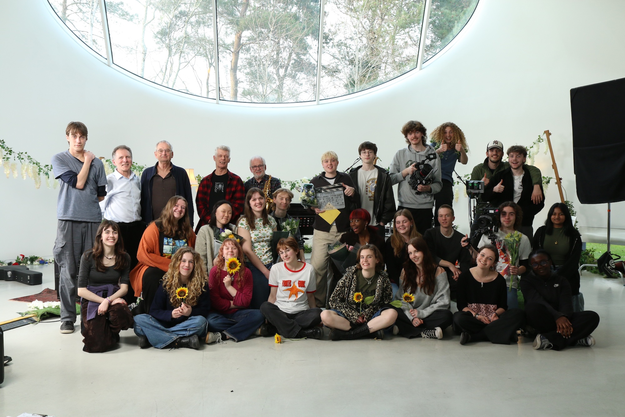 A large group of individuals posing together in a bright space with a circular window. They are arranged in two rows, some holding sunflowers, with greenery and production equipment in the background.