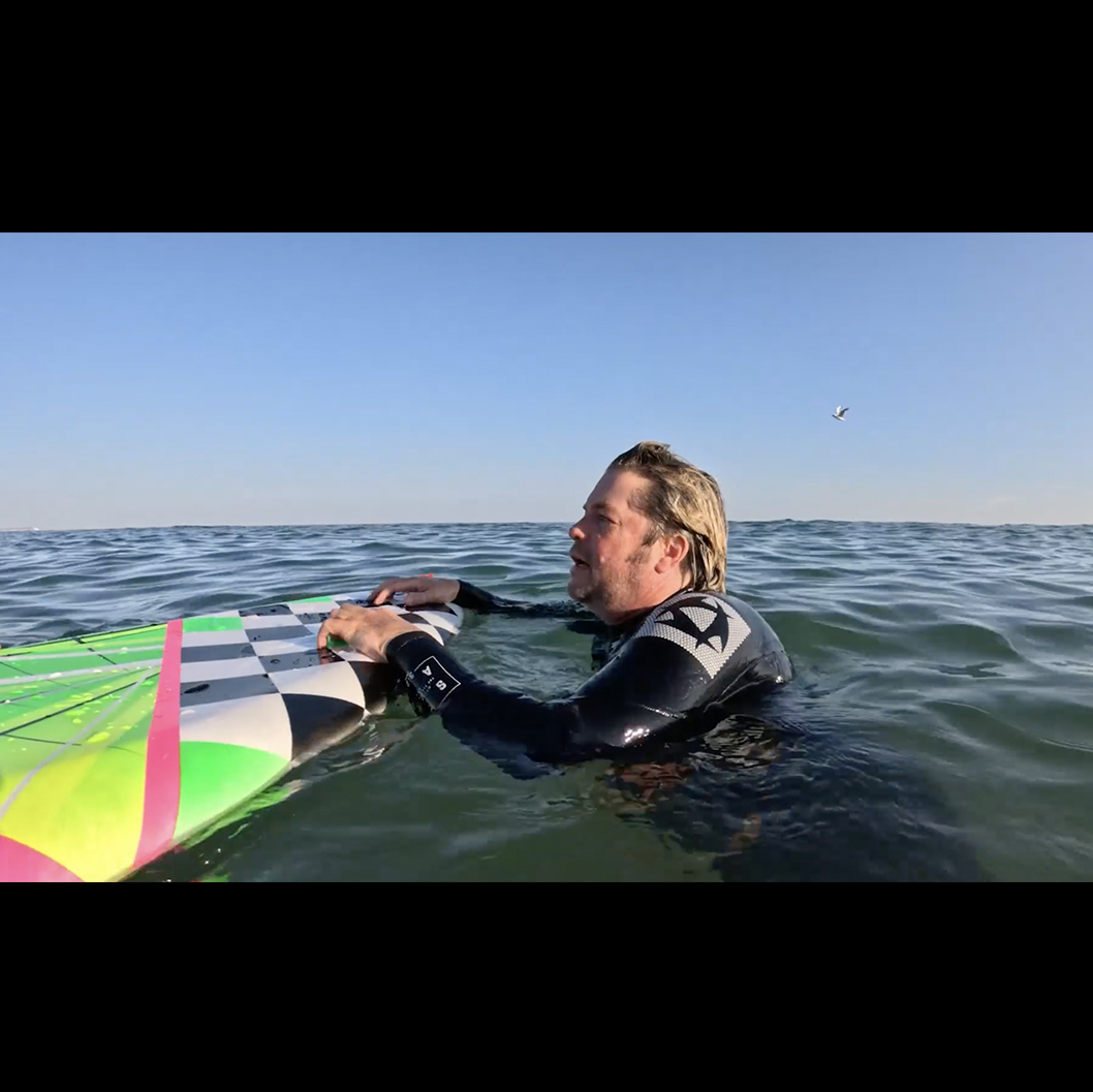 Man in wetsuit floating in ocean water holding green and pink windsurfing sail under blue sky with clouds.