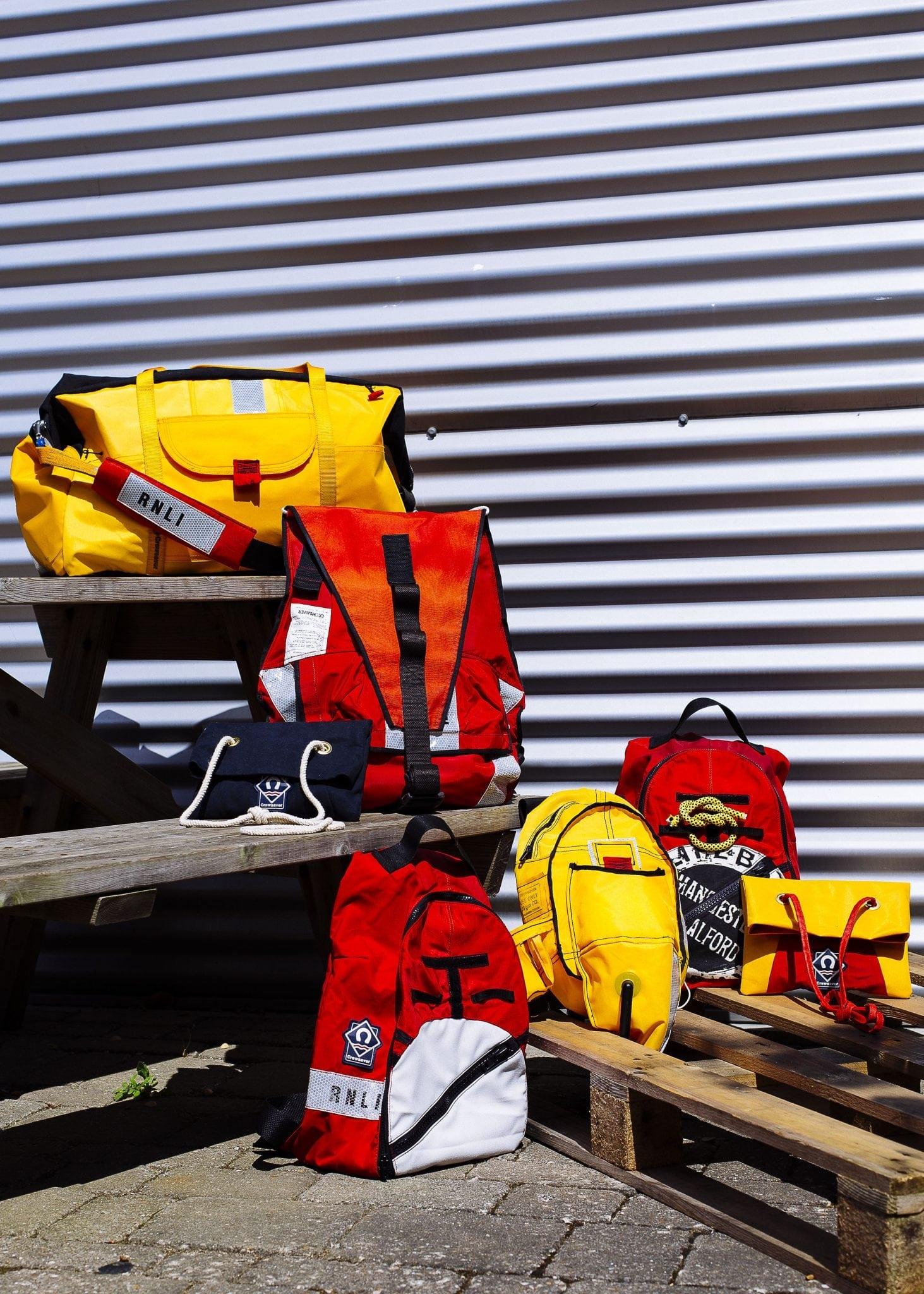 Yellow and red RNLI bags made by BA (Hons) Fashion students sit on benches in front of a large metal door.