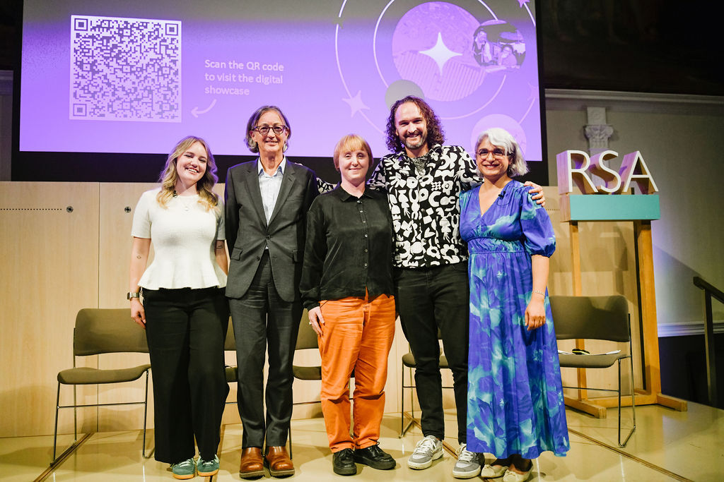 Five people standing together on stage at RSA event, including woman in blue floral dress and man in orange trousers, with purple presentation screen behind.