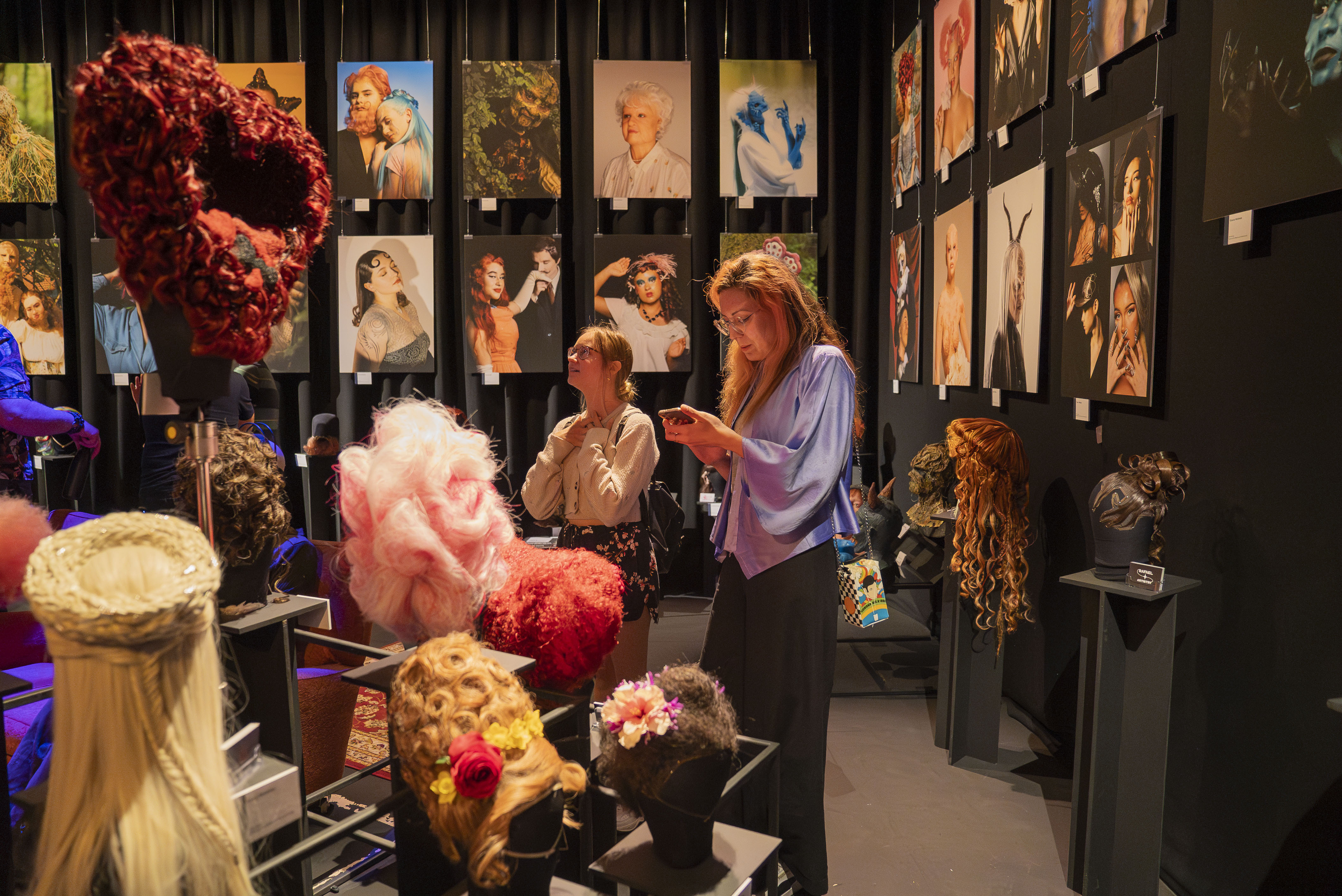 Woman in purple top examining display cases with wigs and masks. Gallery walls covered in framed portraits. Red wreath hangs left.