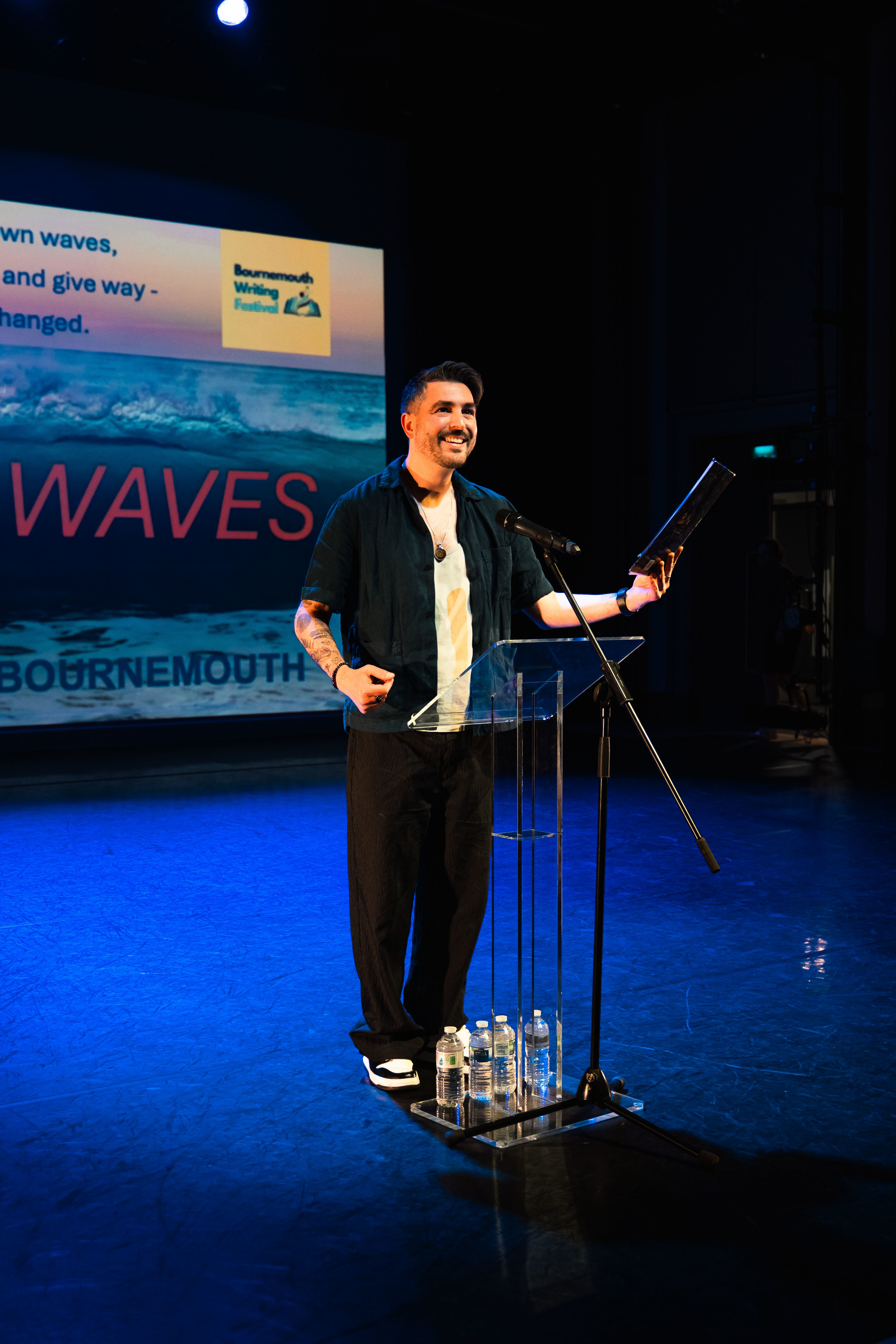 Dr J Cole, course leader of BA (Hons) Creative Writing at Arts University Bournemouth, standing at a lectern on a stage, smiling to a crowd off-camera at Bournemouth Writing Festival.