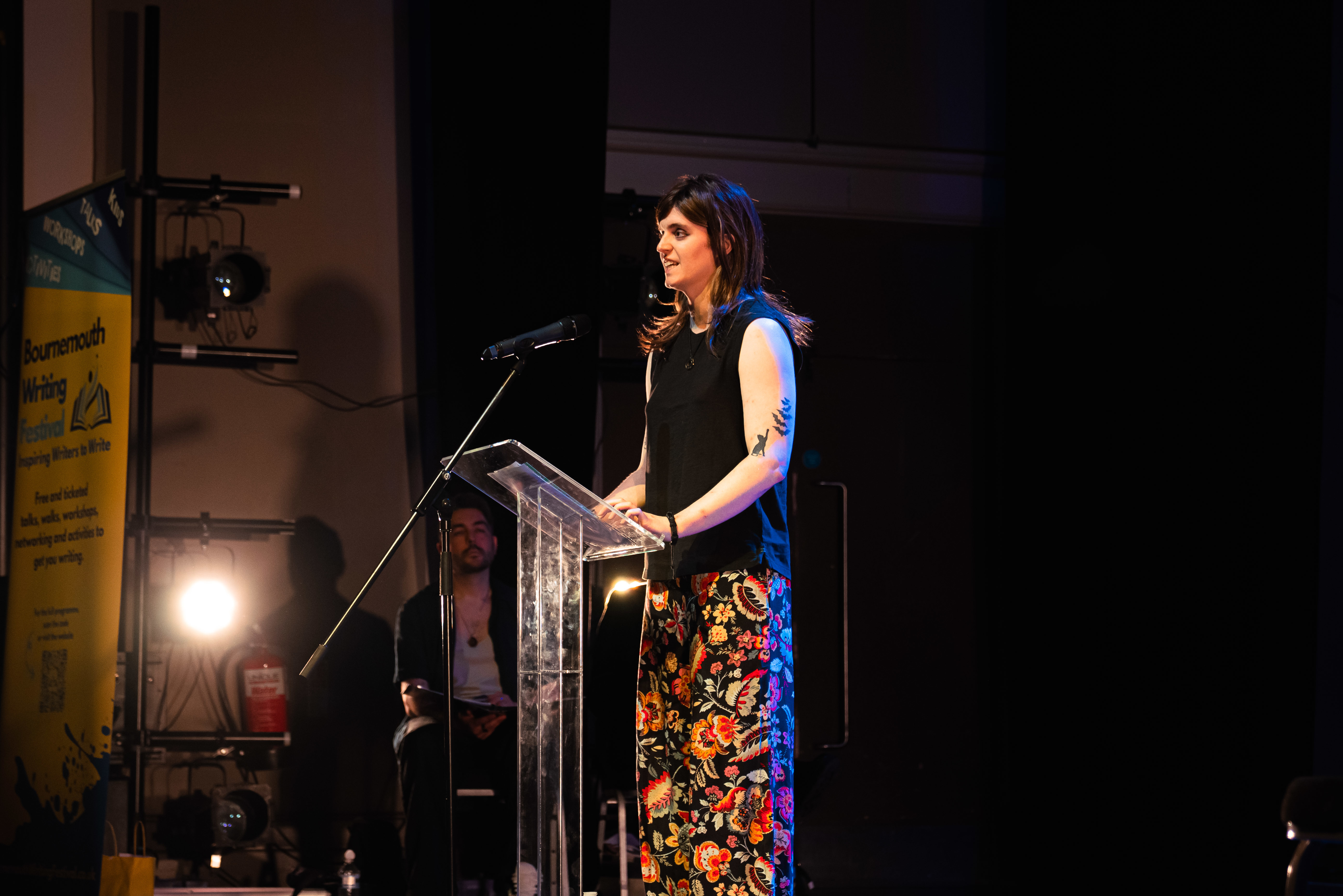 An AUB BA (Hons) Creative Writing student standing at a lectern speaking to an audience off-camera at Bournemouth Writing Festival.