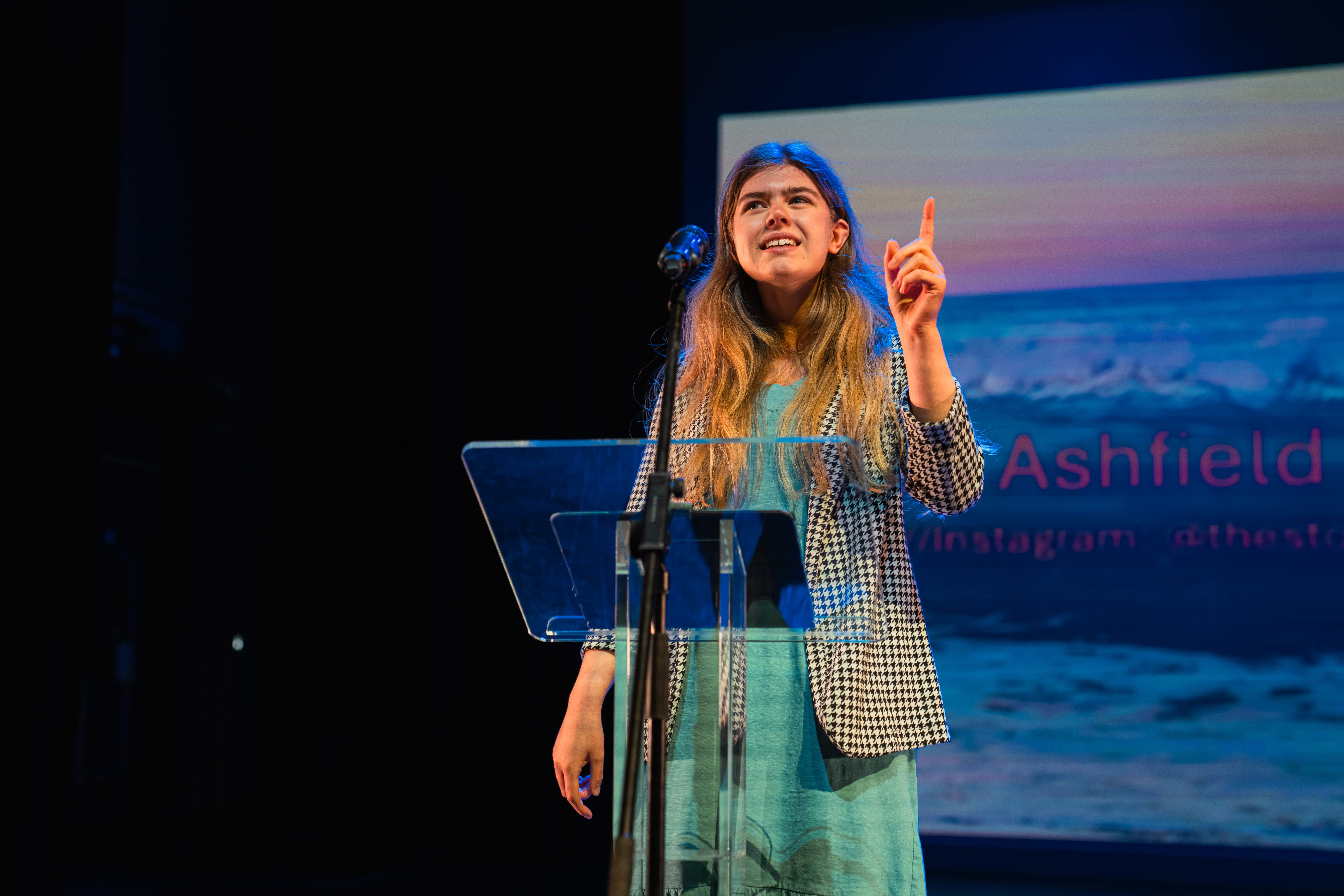 An AUB BA (Hons) Creative Writing student standing at a lectern, pointing to the ceiling while performing for an audience off-camera at Bournemouth Writing Festival.