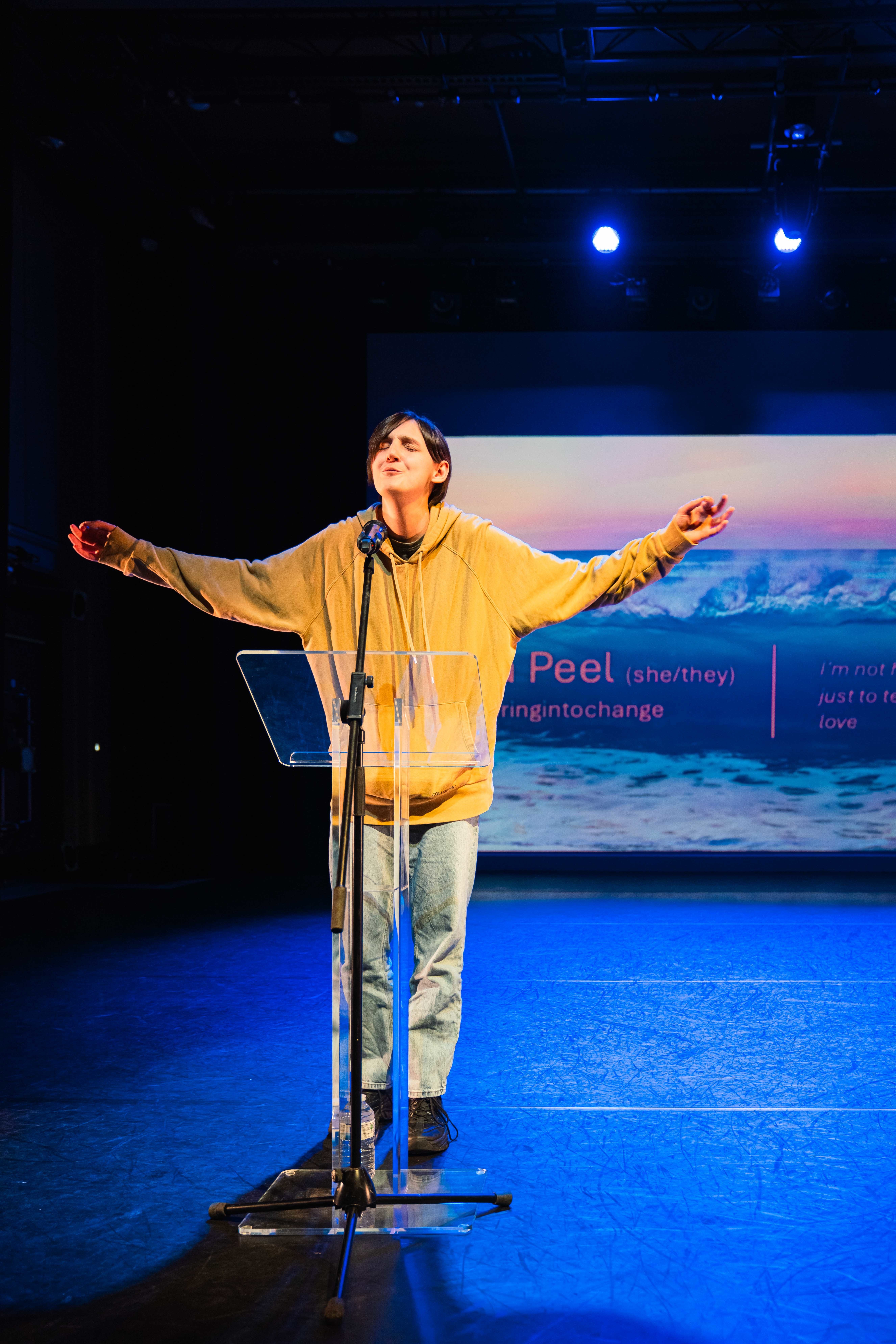 An AUB BA (Hons) Creative Writing student standing at a lectern and speaking into a microphone with arms outstretched, performing for an audience off-camera at Bournemouth Writing Festival.