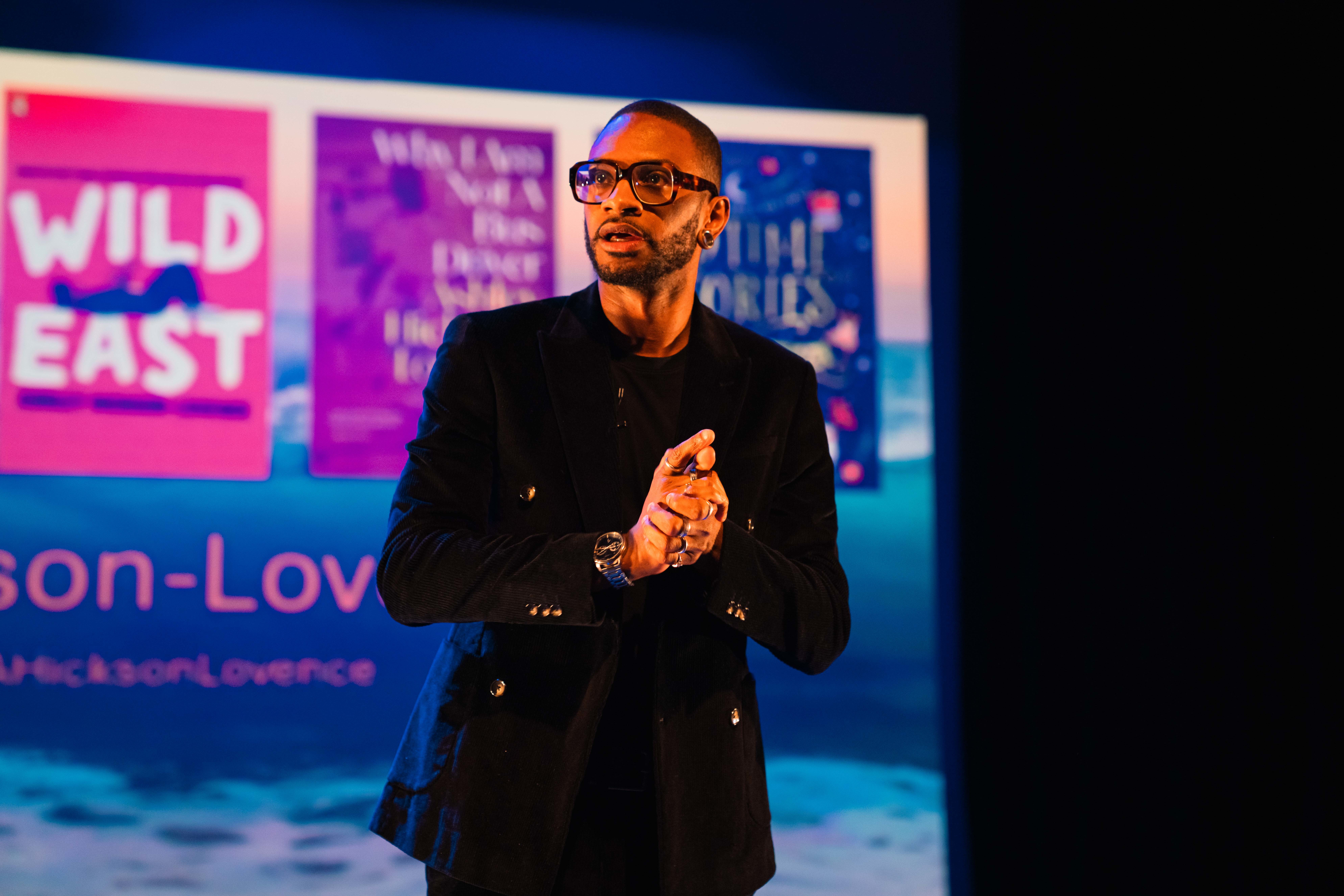 Author A. Hickson-Lovence speaking on stage at Bournemouth Writing Festival, with images of his book covers on a presentation slide behind him.