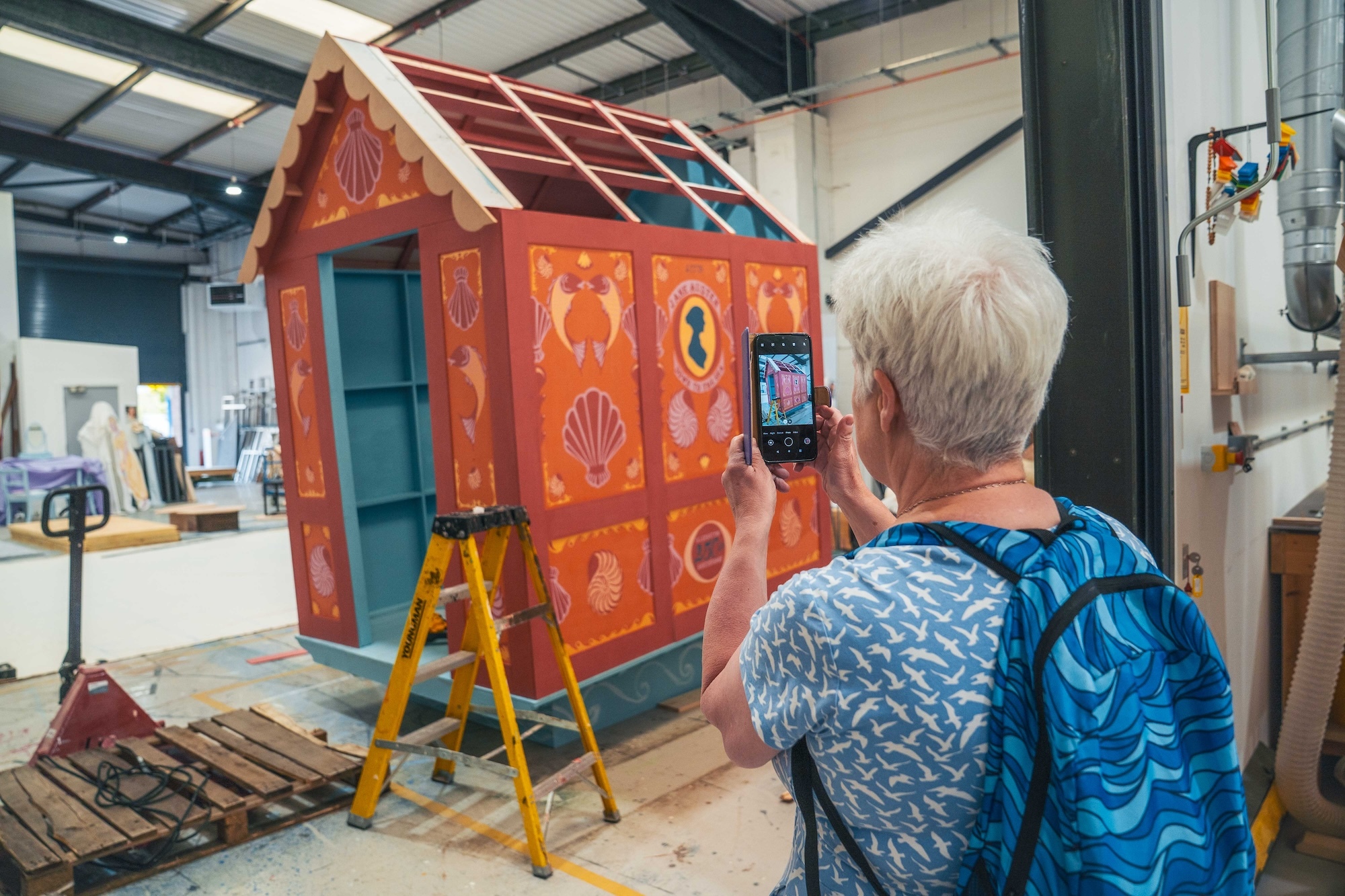 A visitor, viewed from behind, takes a photo on their phone of a small wooden house, painted bright orange in a Regency style, part of a set built at Elliott Road Studios, Arts University Bournemouth.