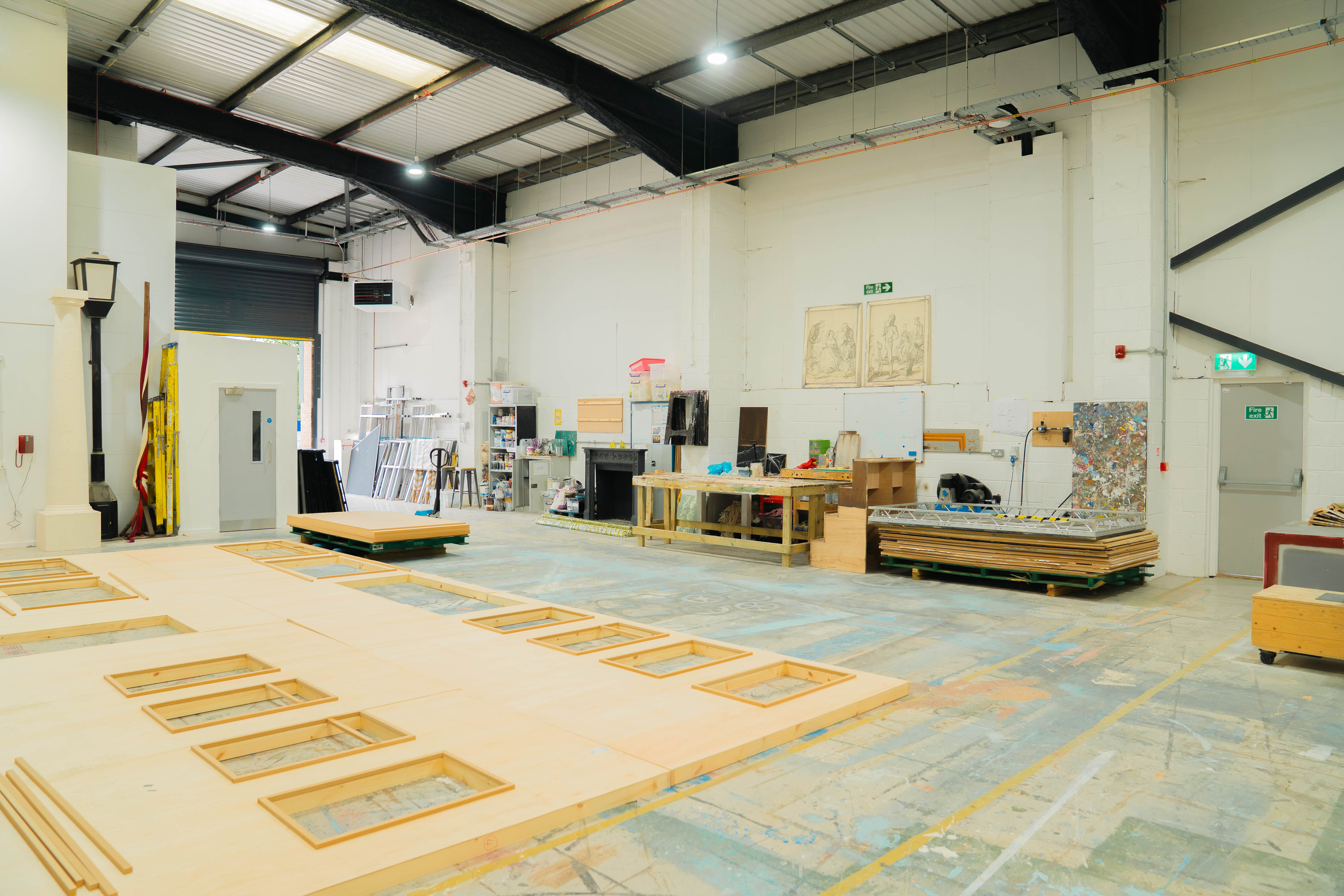 Large warehouse interior with exposed beams, white walls, and concrete floor marked with yellow tape grid lines. Various equipment and materials stored throughout space.