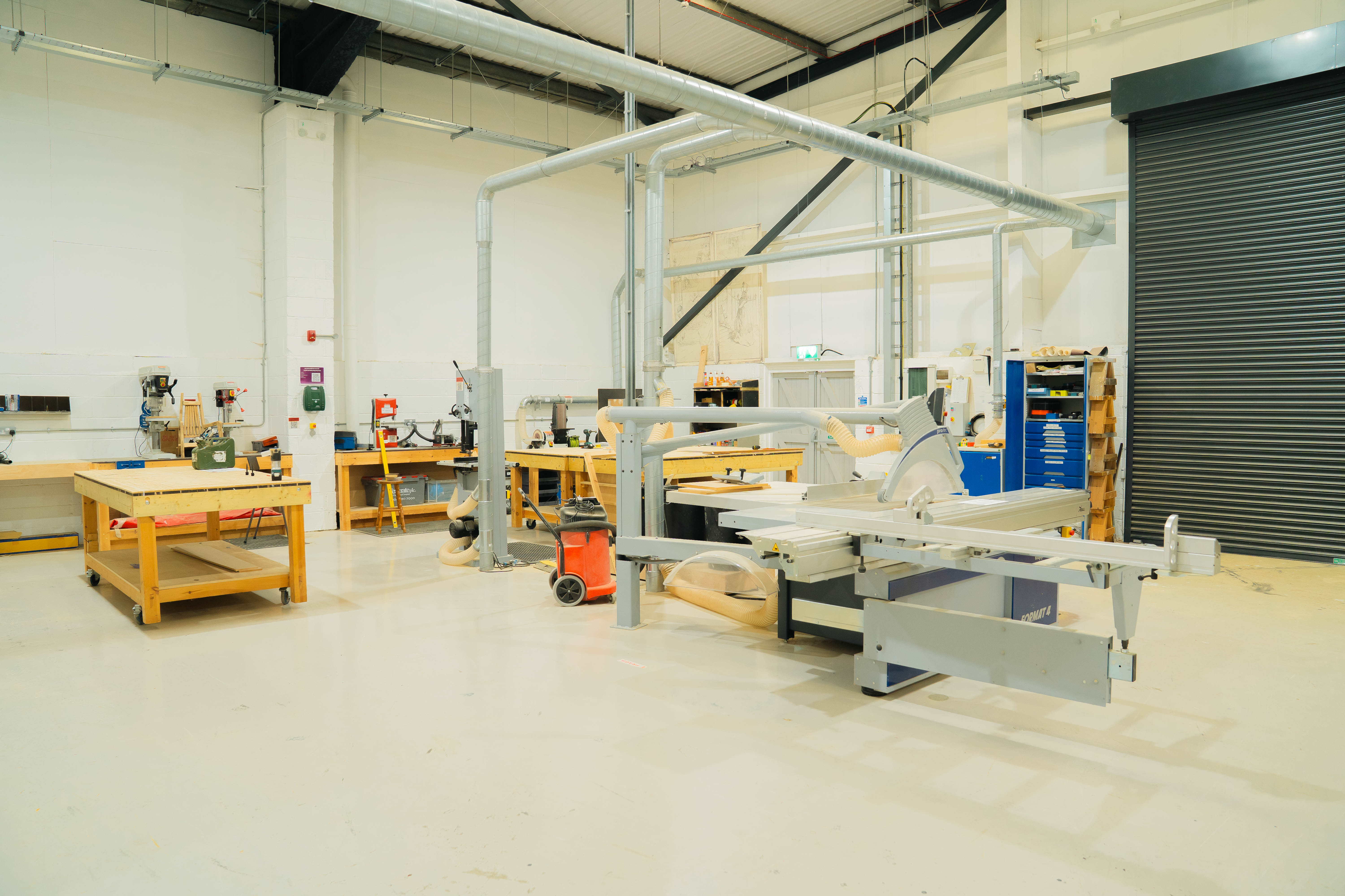 Industrial workshop with white walls, exposed ceiling beams, wooden workbenches, metal equipment, and green roller shutter door.