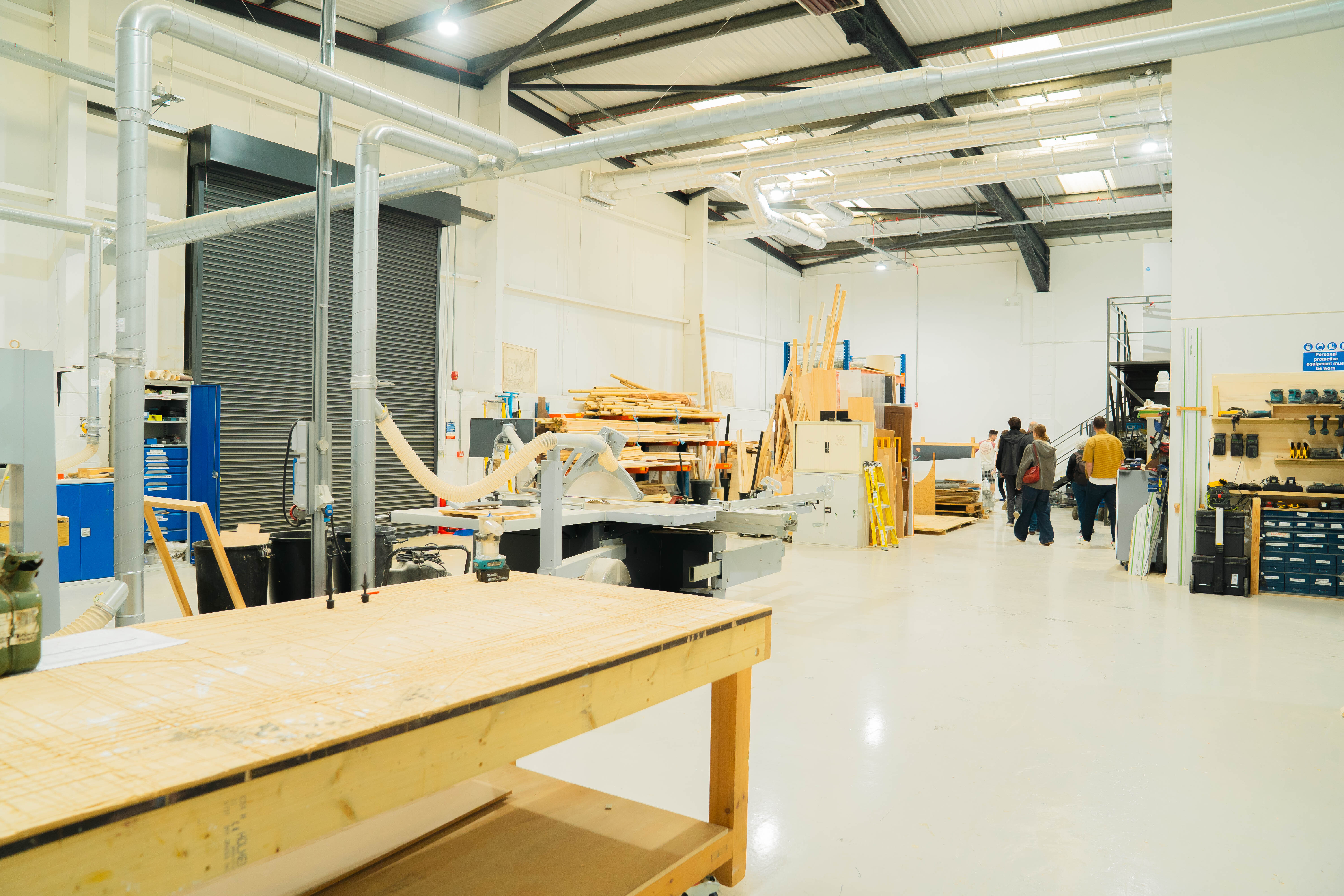 Large workshop interior with wooden workbenches, exposed ceiling beams, white walls, blue machinery, and workers in background.