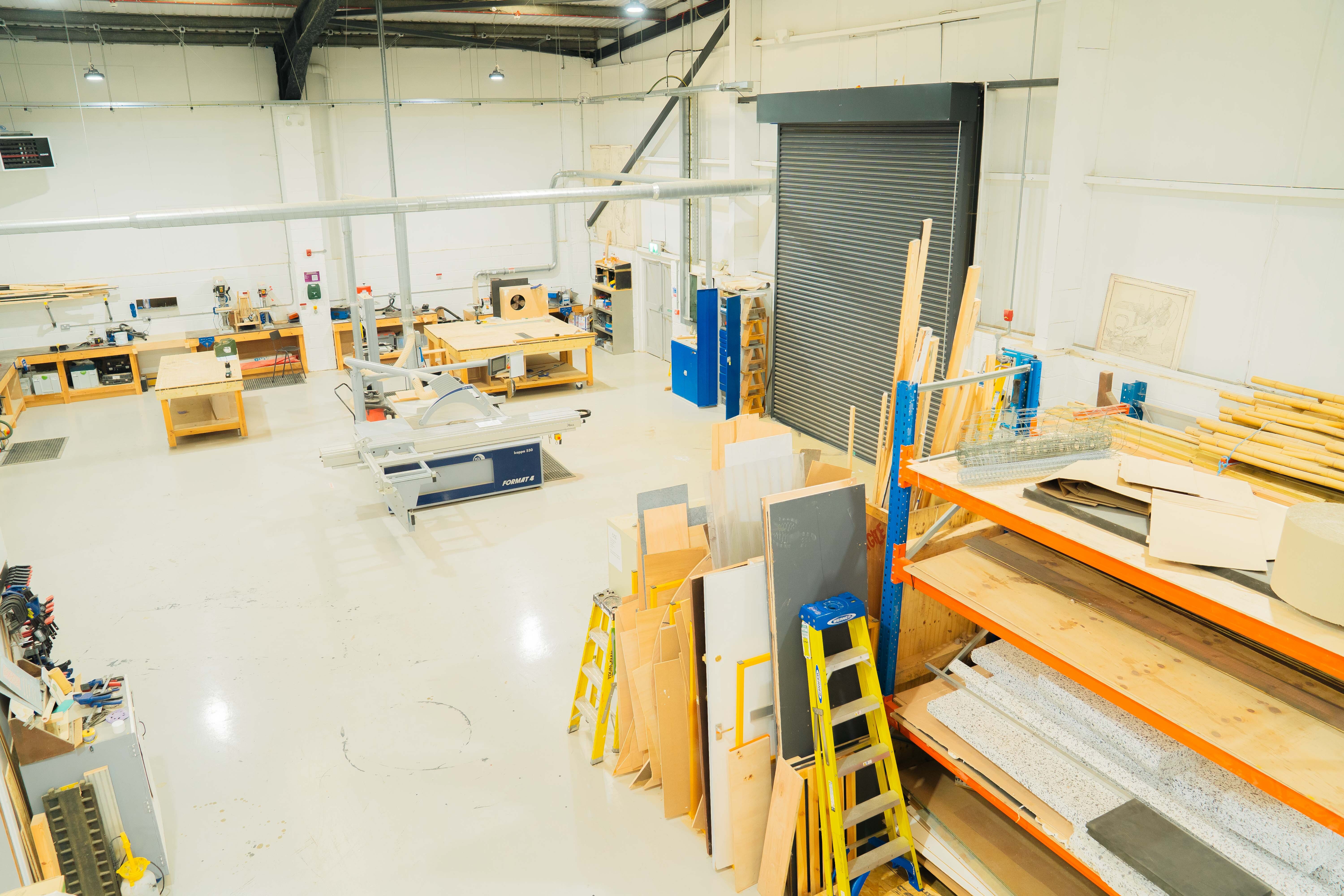 Large warehouse interior with orange shelving units, yellow ladders, cardboard boxes, and metal roller shutter doors on white walls.