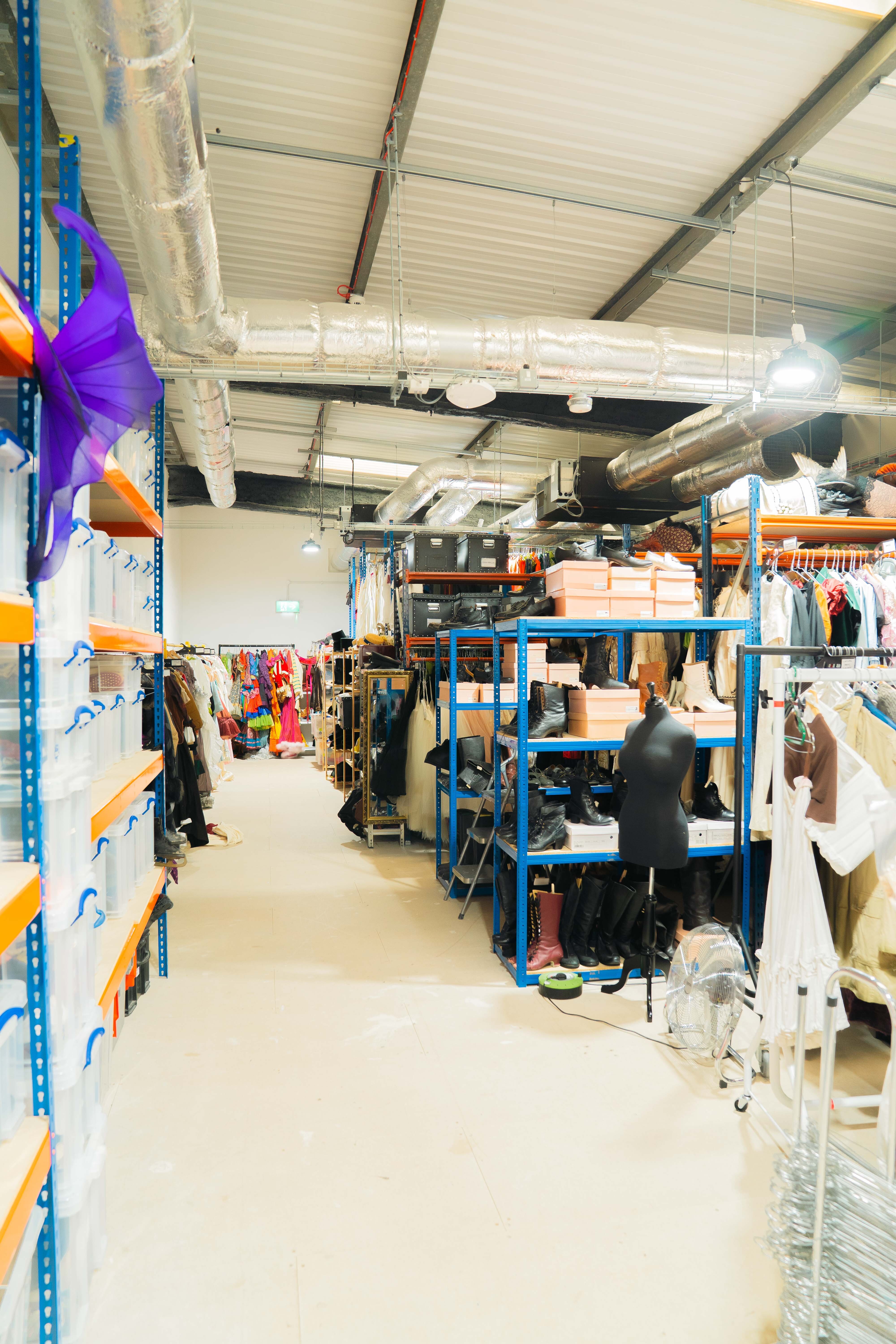 Warehouse interior with blue metal shelving units, concrete floor, white ceiling with exposed beams, and person in dark clothing.