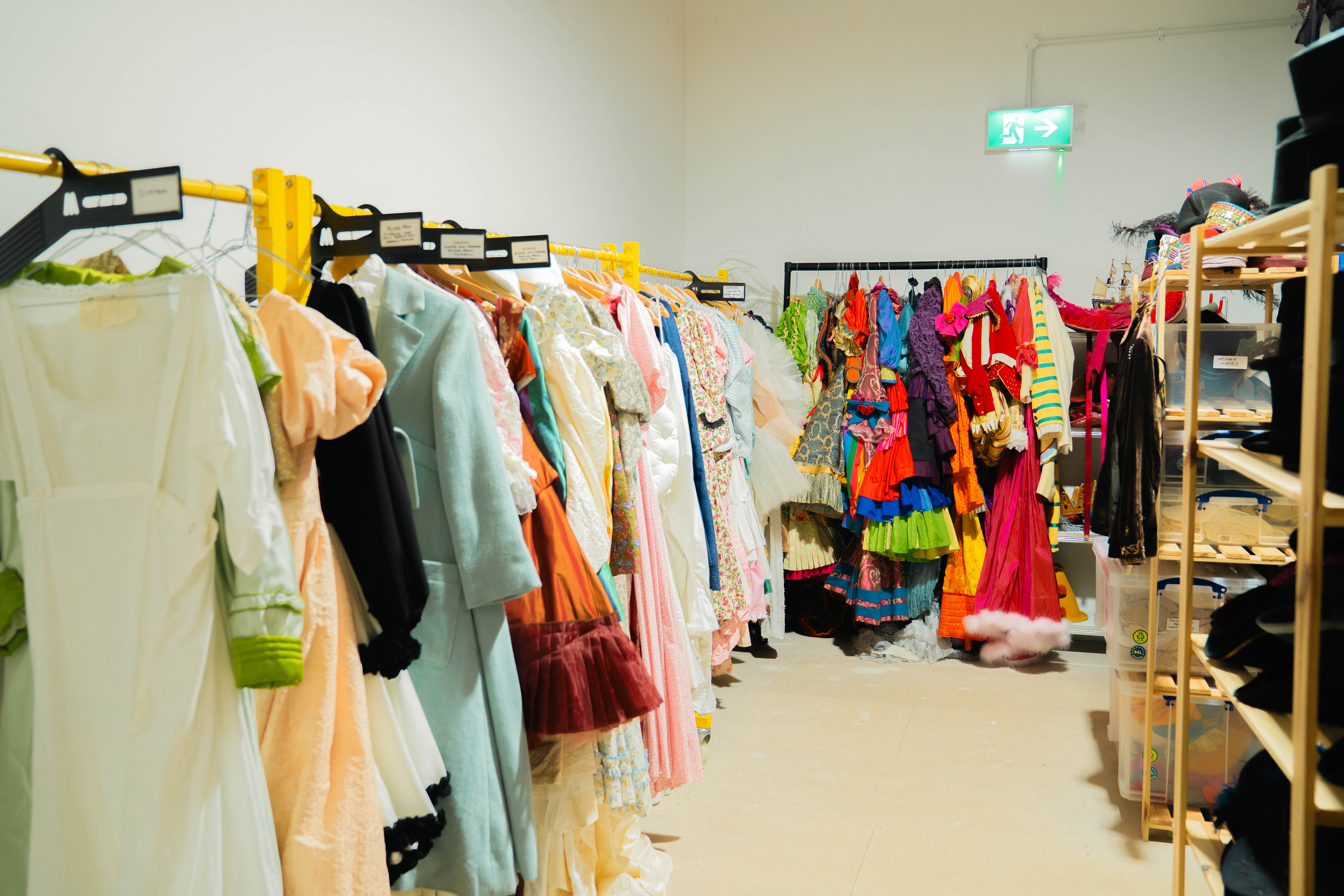 Clothing shop interior with garments on metal rails, including pastel and bright coloured dresses, wooden shelving units, white walls.