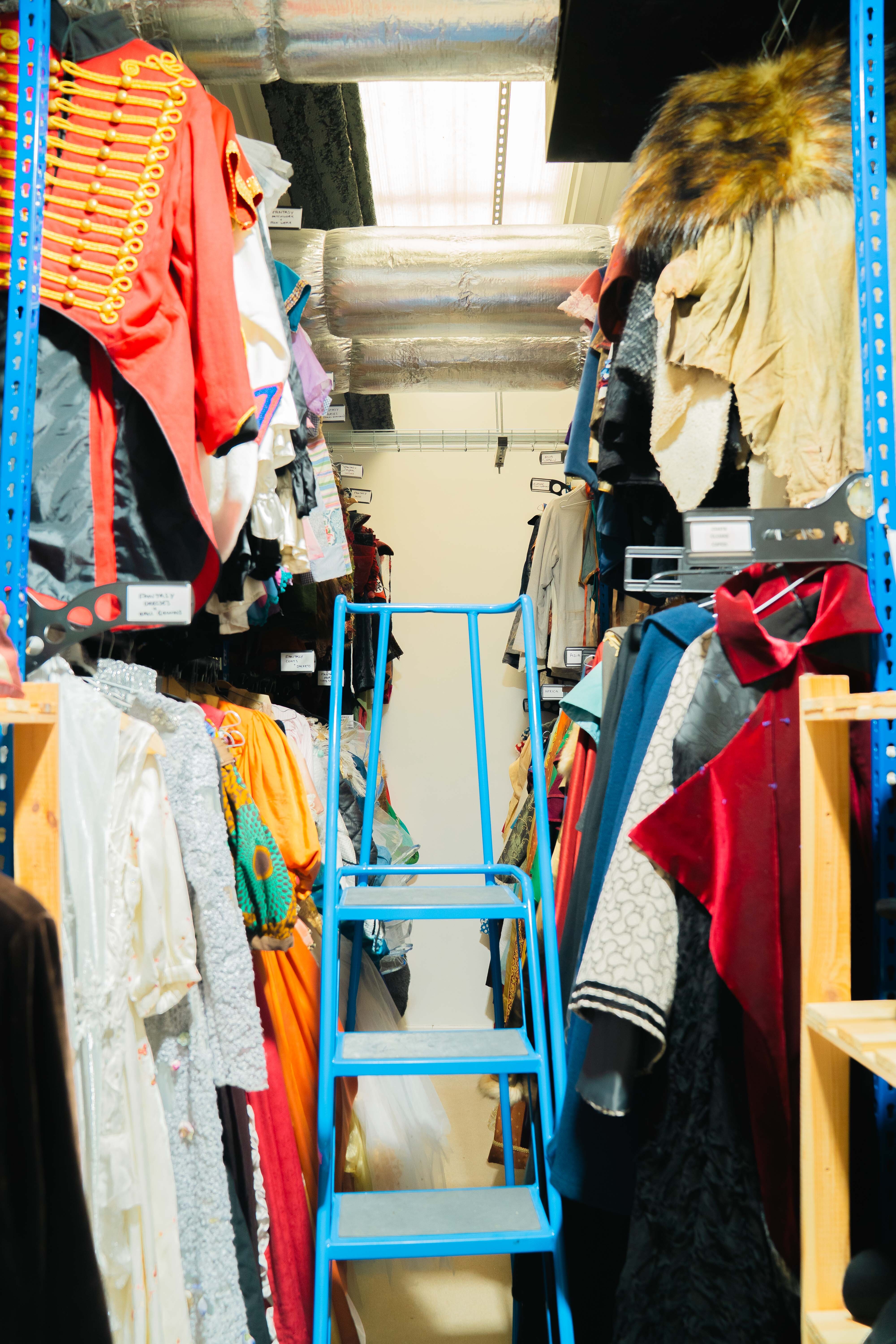 Storage room with blue metal shelving holding various costumes including red military-style jacket, feathered headdress, and colourful garments.