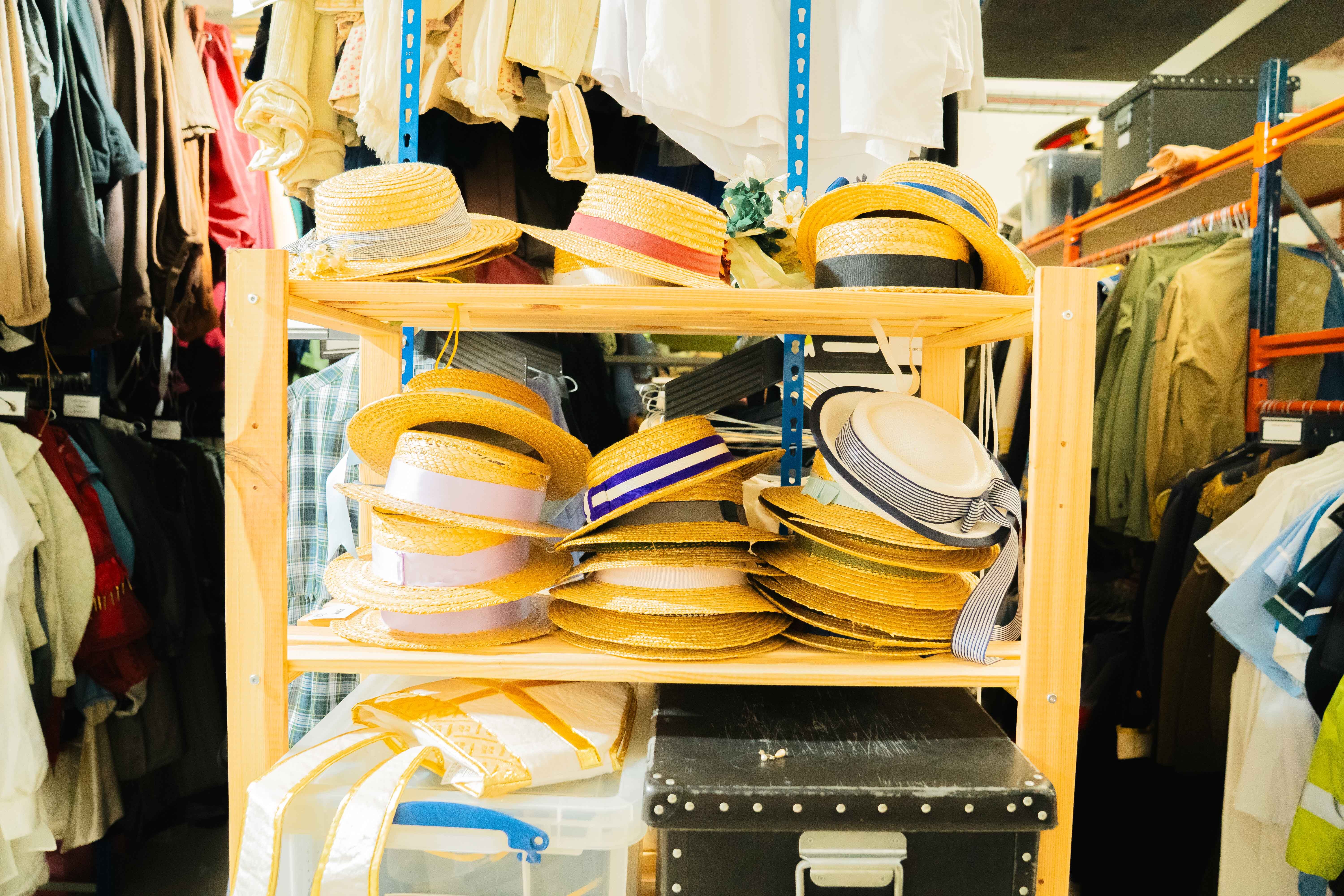 Wooden shelving unit displaying multiple yellow and white striped hats on two levels, with clothing and storage boxes visible around it.