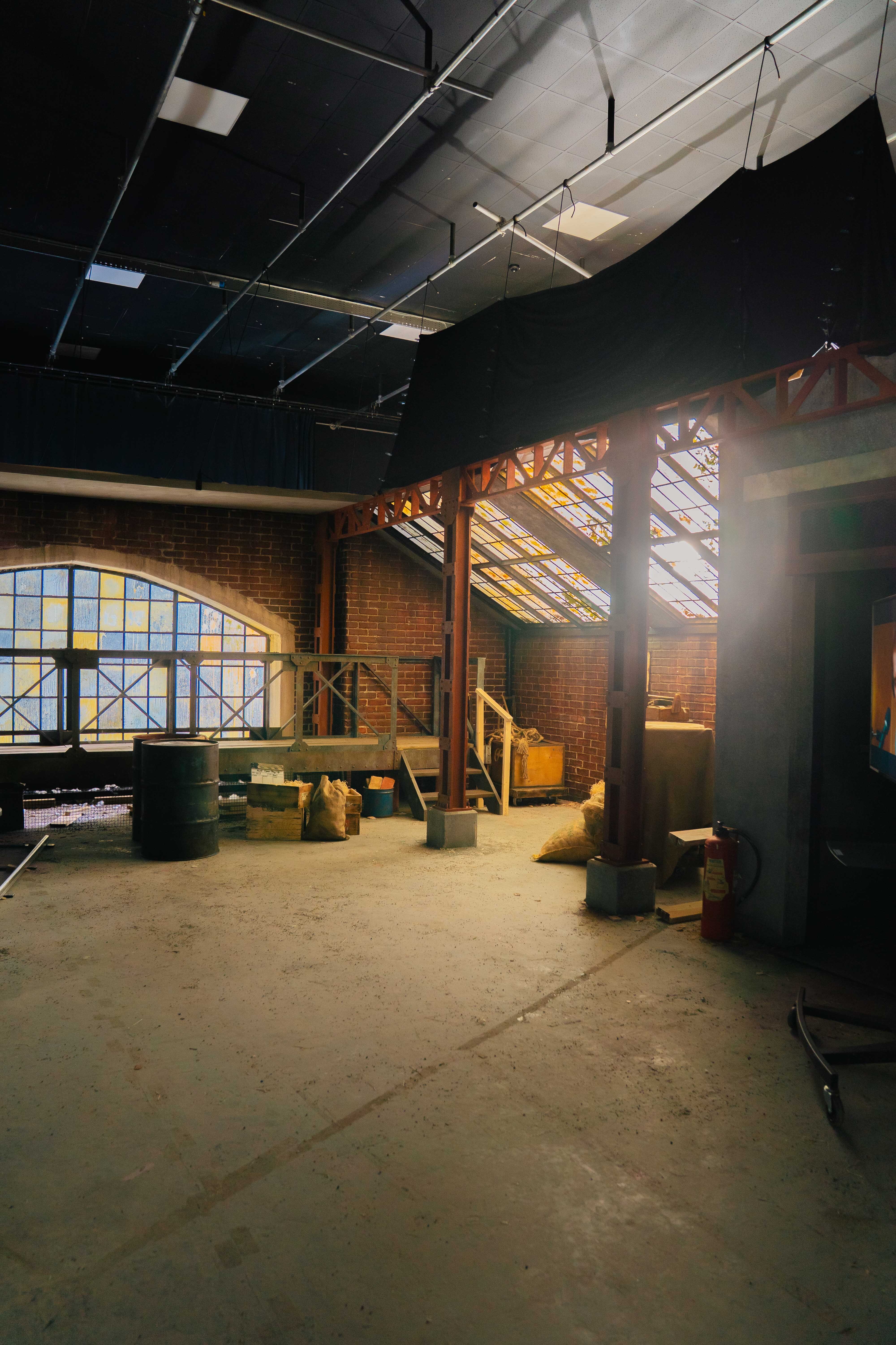 Industrial interior under renovation with exposed brick walls, damaged ceiling tiles, concrete floor, and sunlight streaming through skylights.