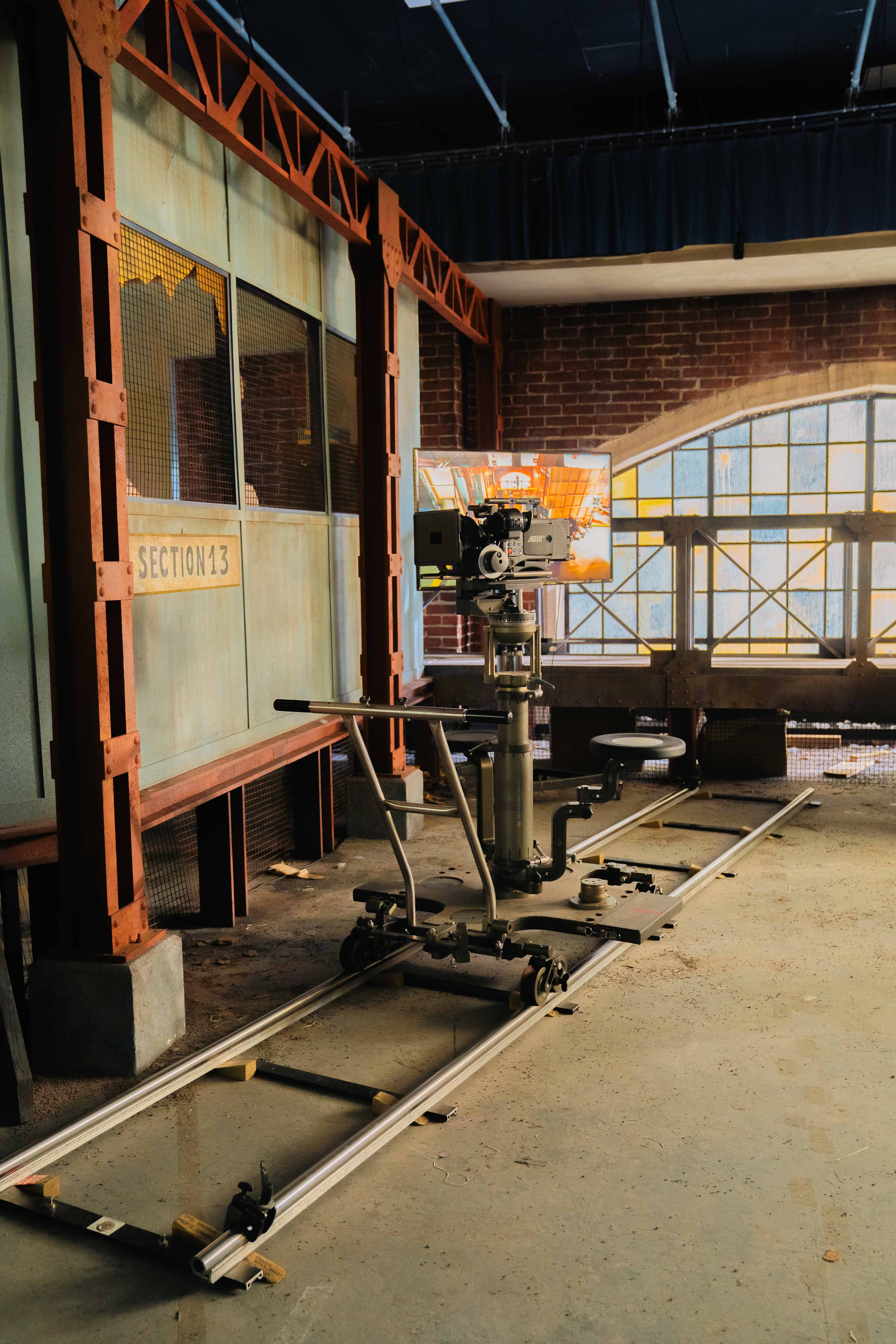Television studio set with brick walls, large windows, camera equipment on tracks, and "Restrooms" sign visible on left wall.