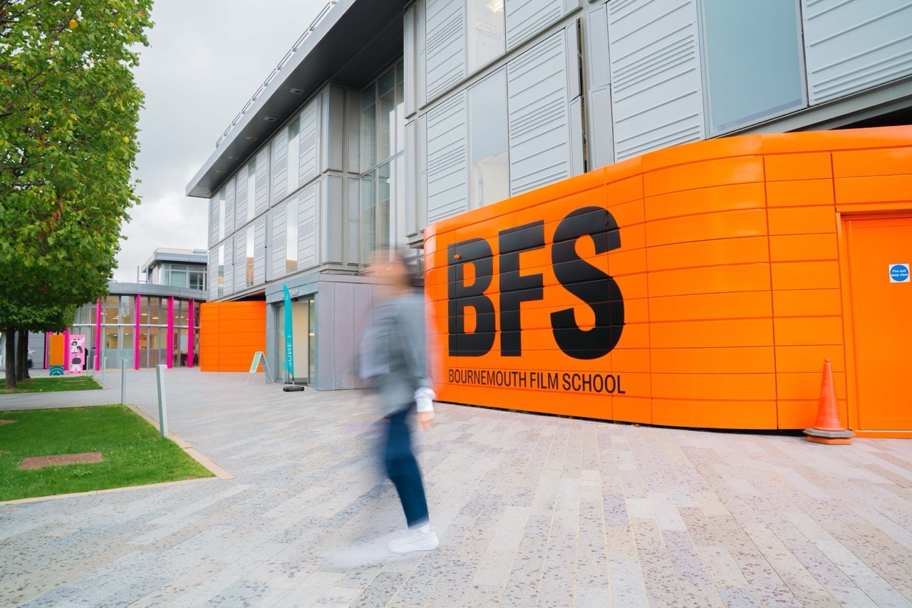 Modern building with bright orange facade displaying "BFS" text. Blurred figure walking past colourful entrance area with green trees.