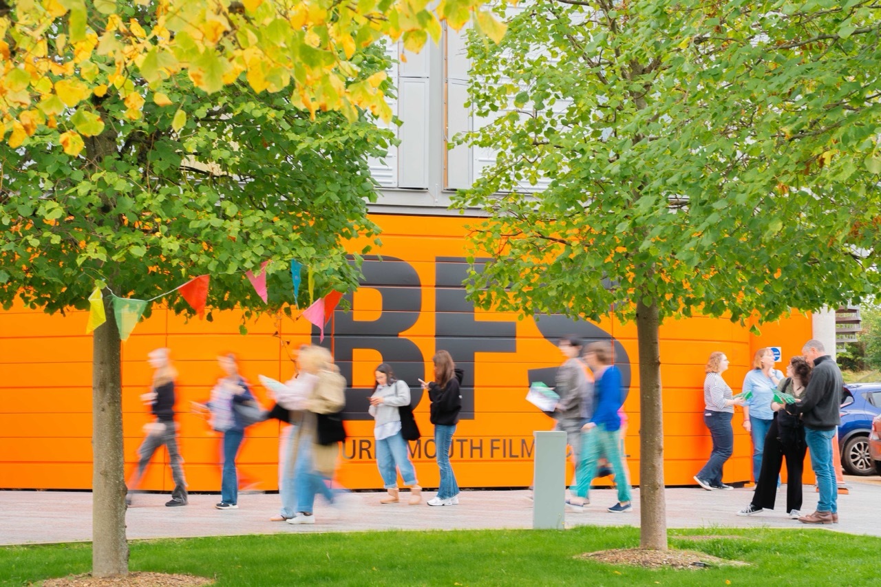 People queuing outside orange building with "BFI" signage, framed by green trees with yellow autumn leaves.