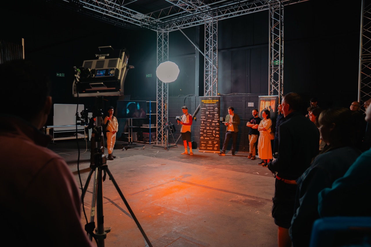 Film set with metal scaffolding, orange-lit stage area, crew in hi-vis jackets, camera on tripod in foreground, dark studio environment.