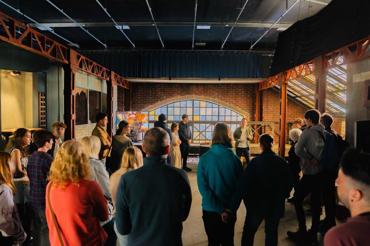 Group of people standing in industrial brick building with exposed ceiling beams, large arched window, and warm lighting.