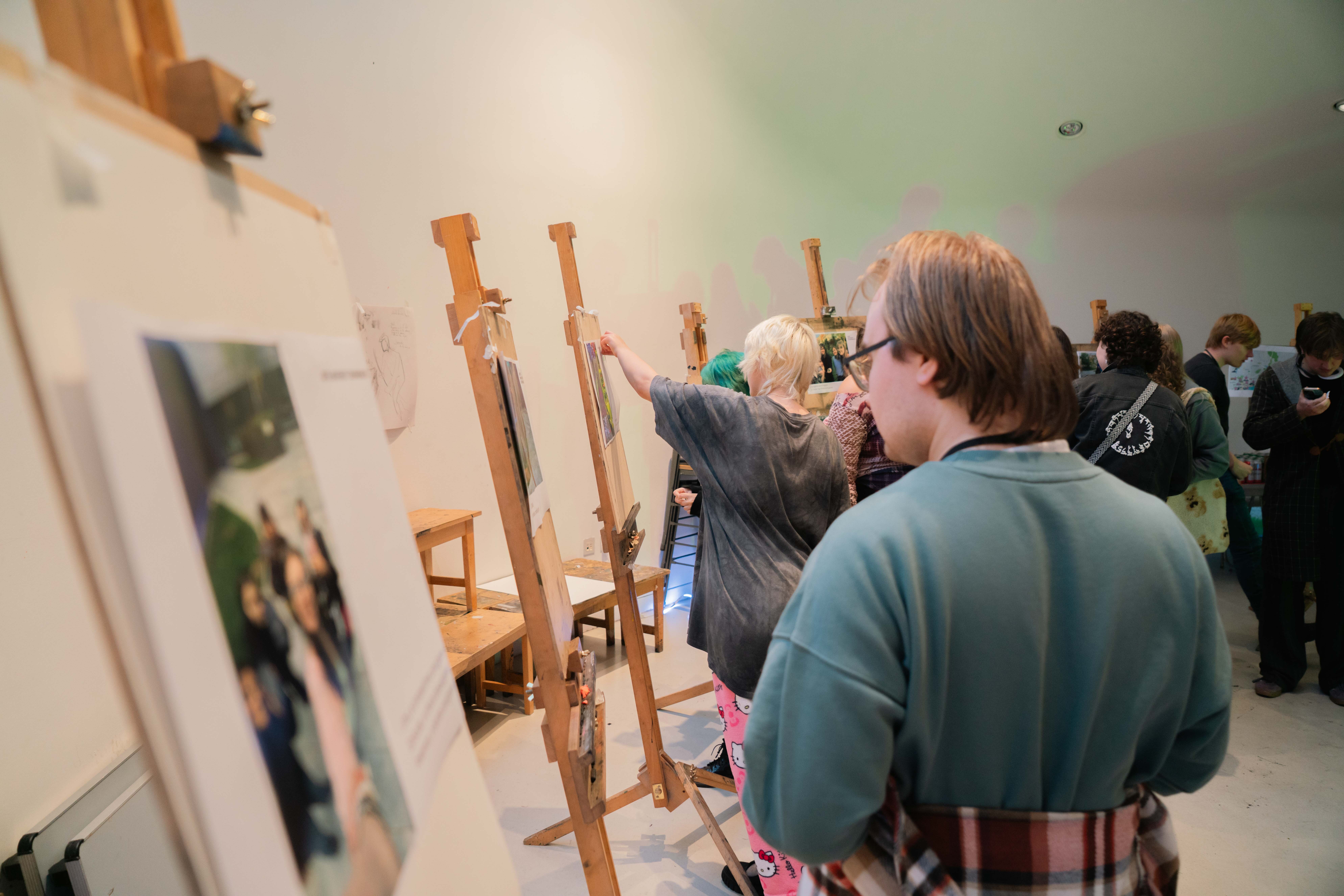 Woman in grey jumper painting at wooden easel in art class with other students working at easels in background.