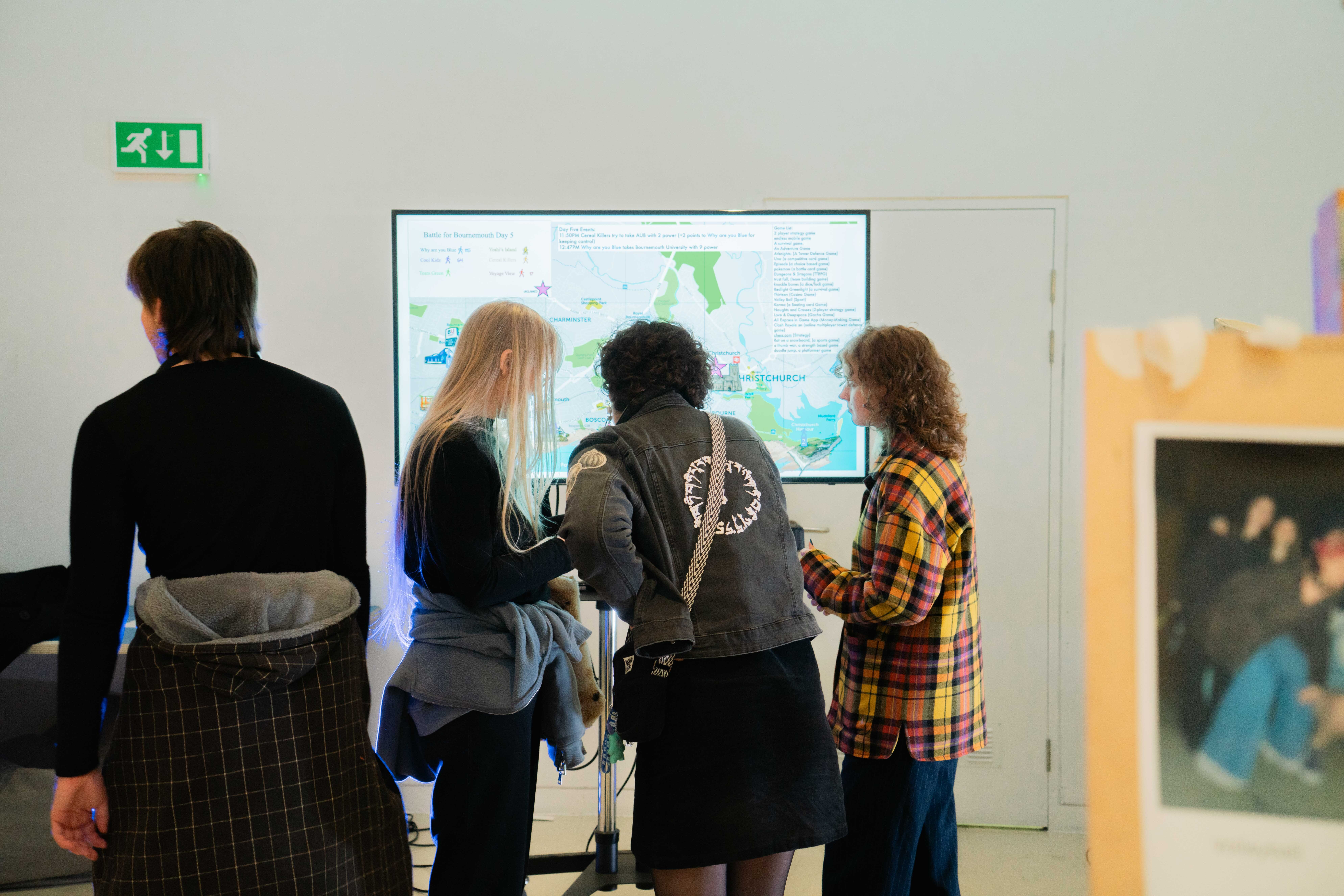 Four people standing before large wall-mounted display screen showing map with green and blue areas. Green exit sign visible upper left.