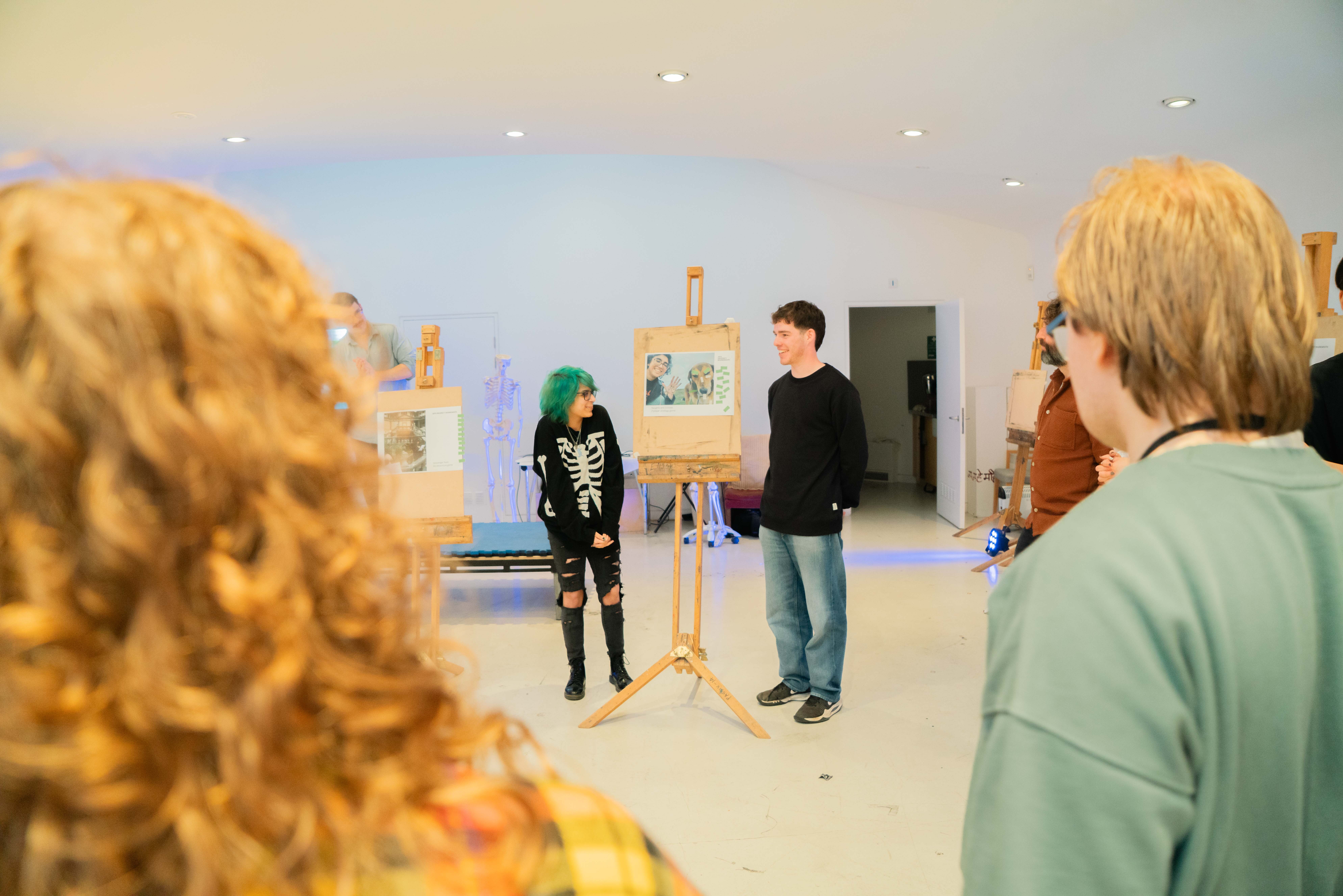 Group of people in white gallery space with paintings on easels, woman with green hair in skeleton top, man in dark jumper speaking.