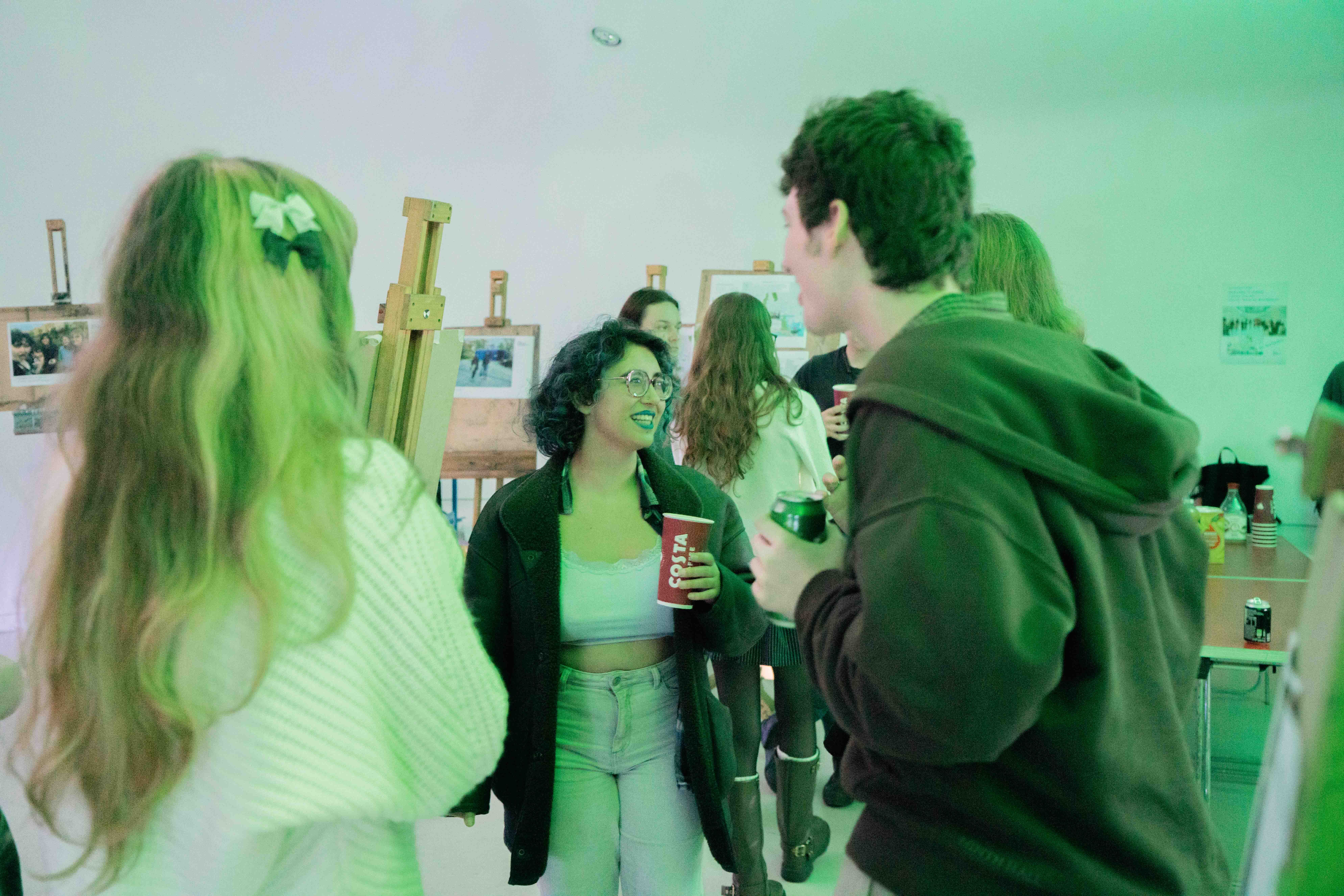 People in green costumes and clothing gathered indoors with green-tinted lighting, person on left wearing white with long blonde hair.