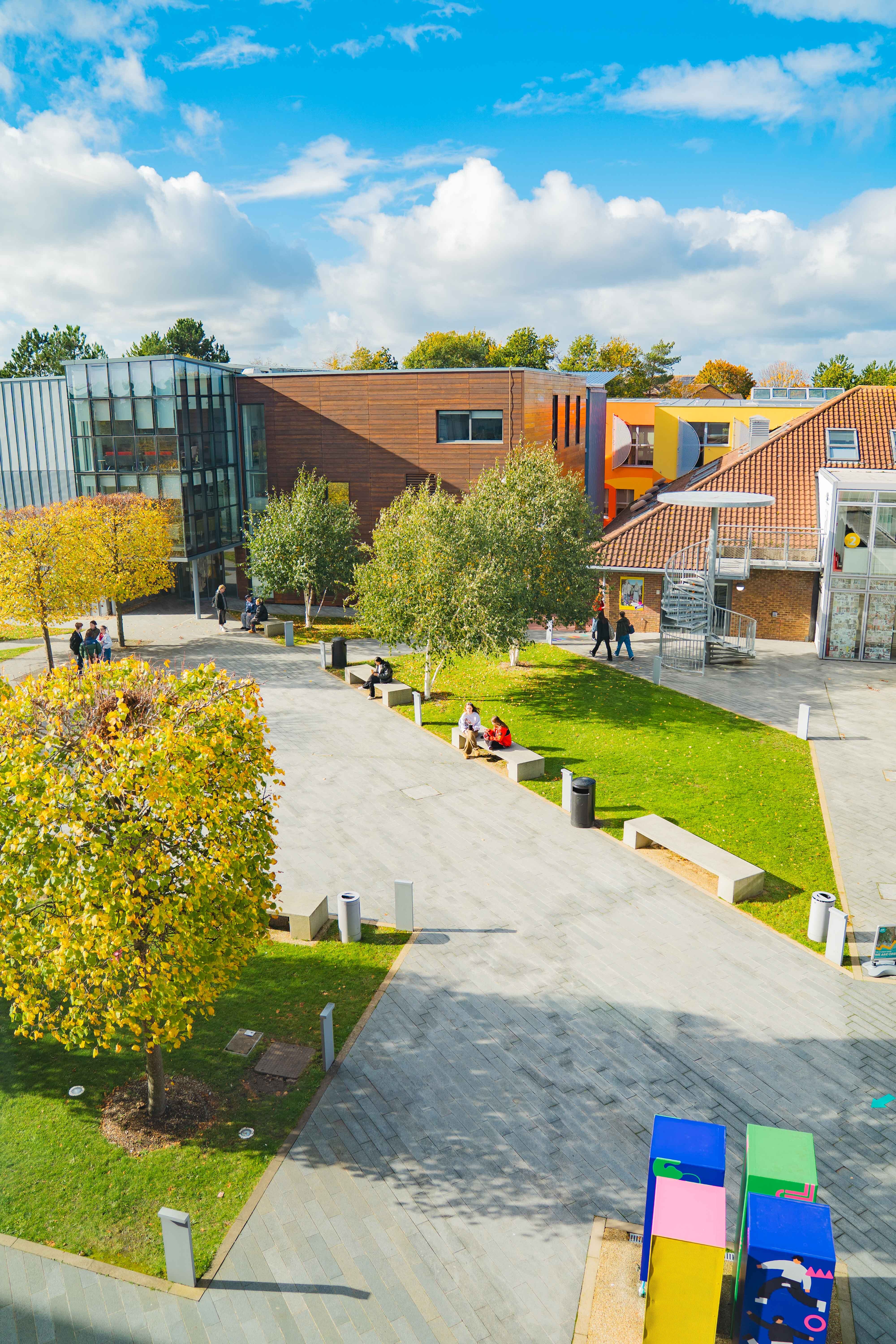 Arts University Bournemouth campus with glass buildings, orange and red-tiled structures, green lawns, paved walkways, and colourful play equipment.