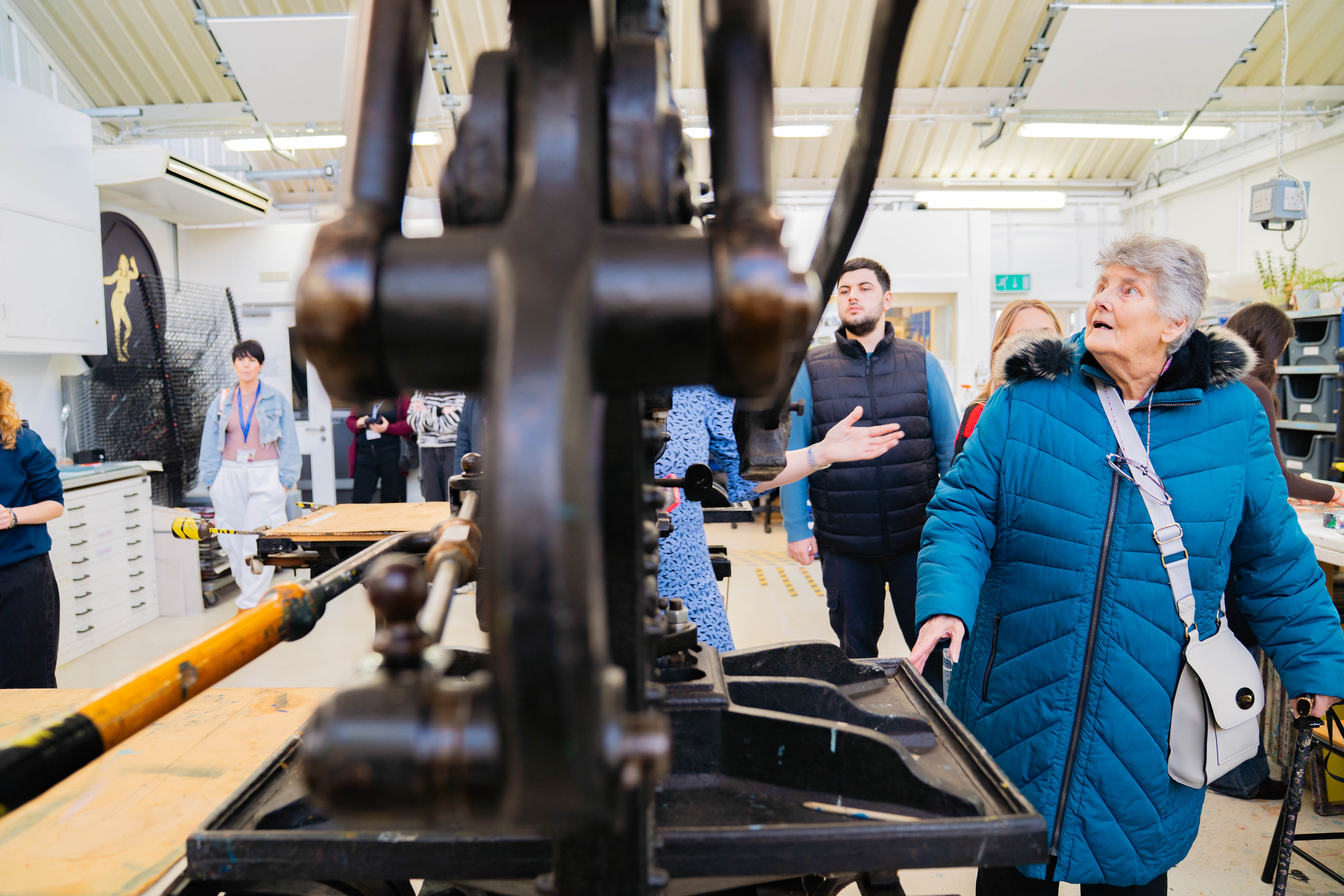 Elderly woman in blue coat observes large black printing press machine in workshop with other visitors and staff in background.