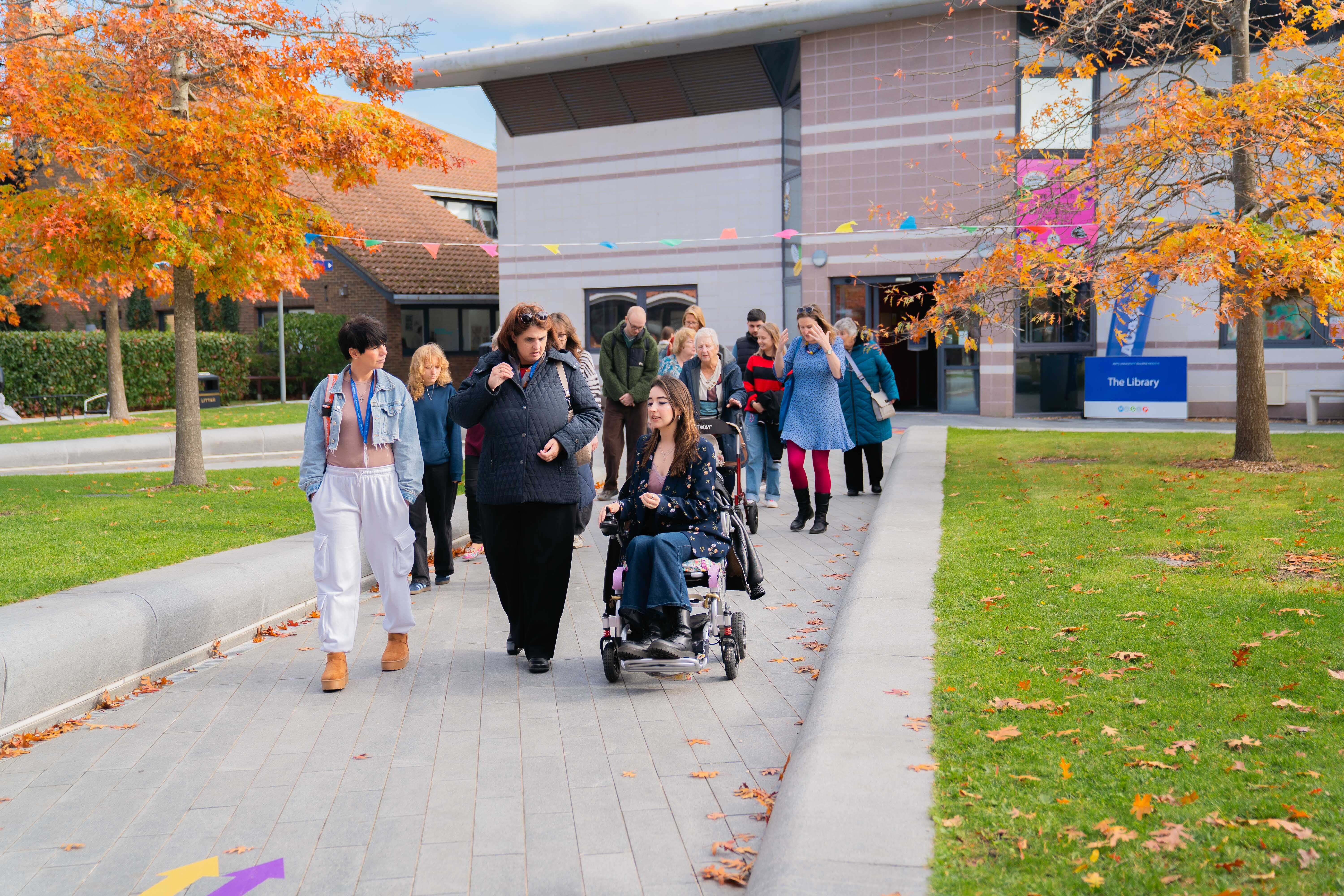 Group of people walking on pavement towards modern building, with person in wheelchair. Orange autumn trees frame concrete structure.