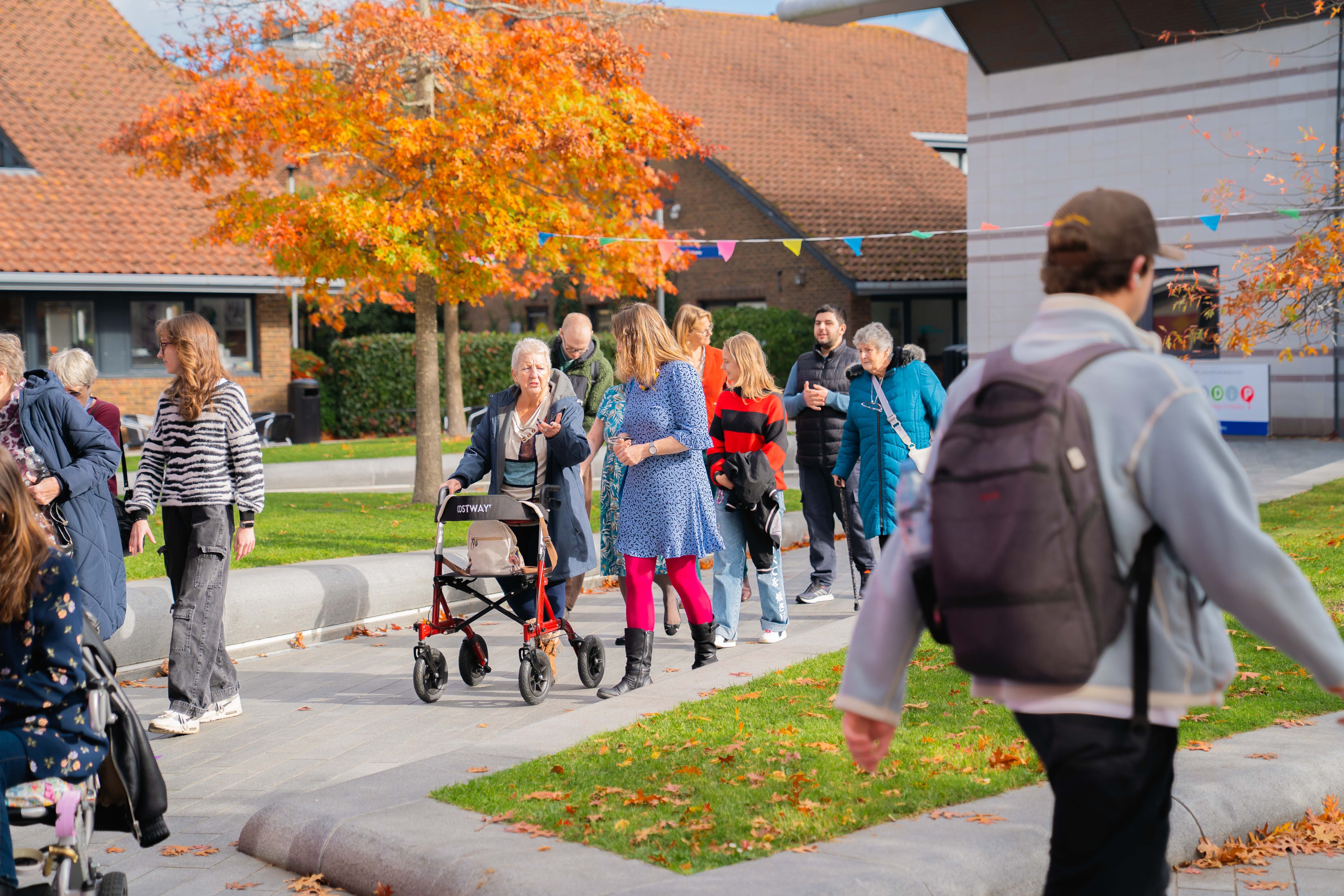 Group of people gathered on pavement near building with orange autumn tree and green lawn, man with rucksack in foreground.