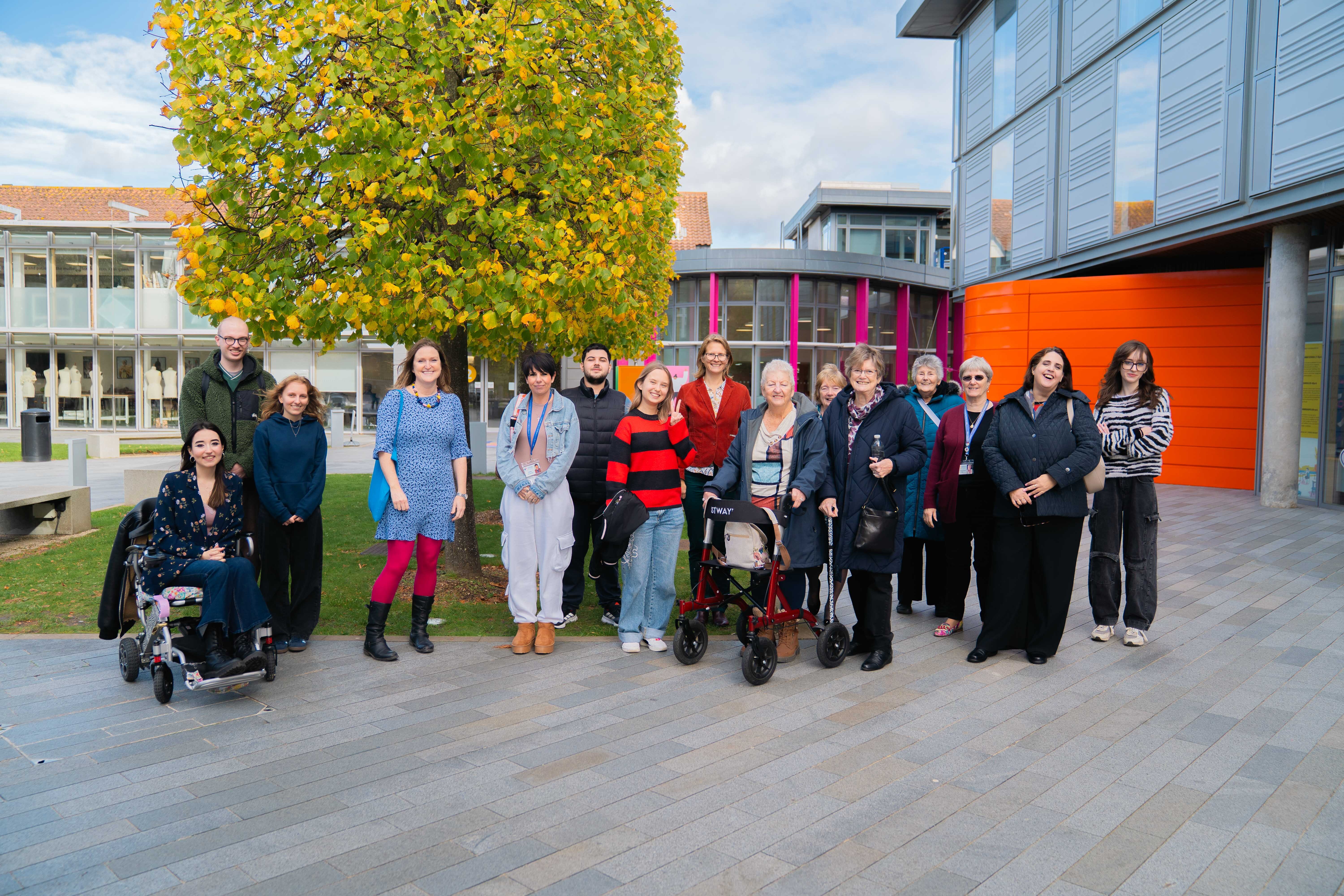 Group of people with mobility aids standing outside modern buildings with orange and purple facades, large tree in background.