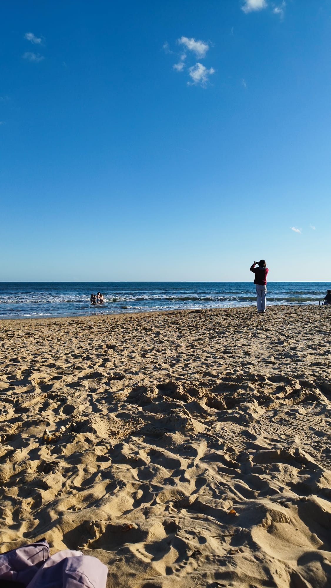 Portrait shot of Bournemouth beach, sand and a bright blue sky with people swimming and someone stood looking out to the ocean.