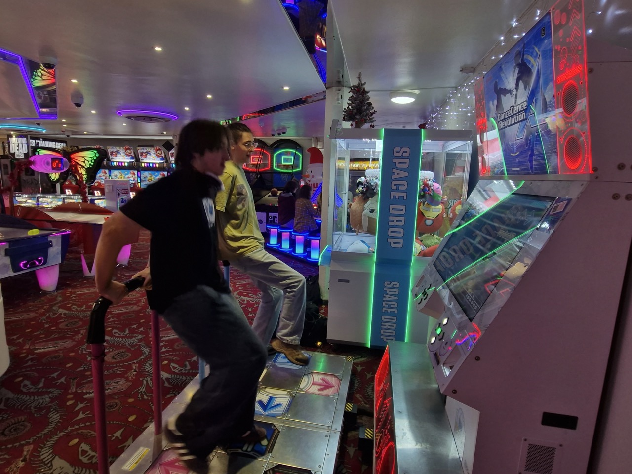 Two students holding onto a rail on a DDR (Dance Dance Revolution) machine, colourful arcade neon lights in the background behind them.