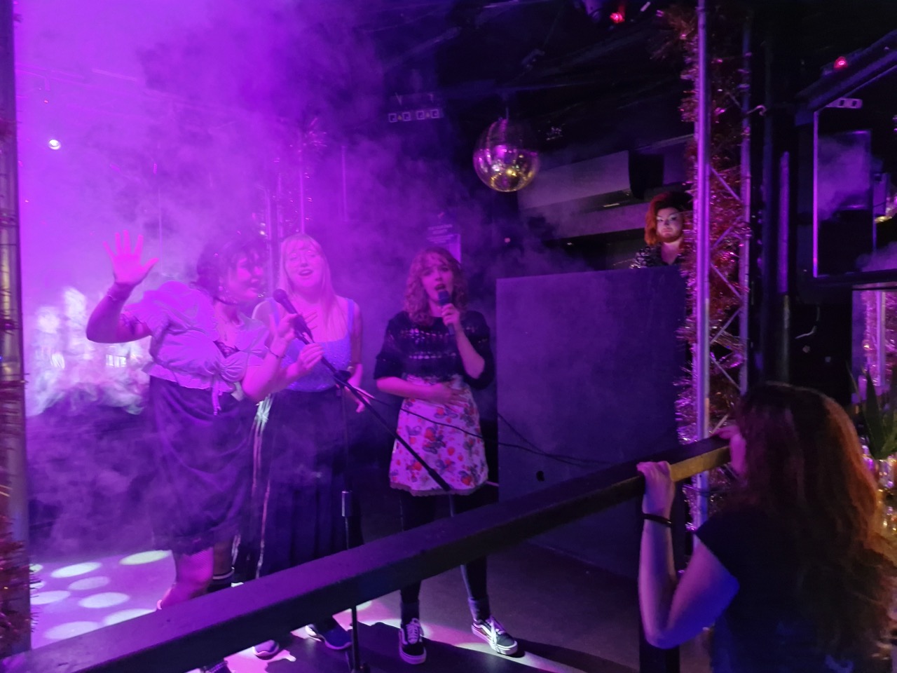 A group of three students singing karaoke at DYMK, purple lighting and smoke, drag queen DJ'ing in the background.