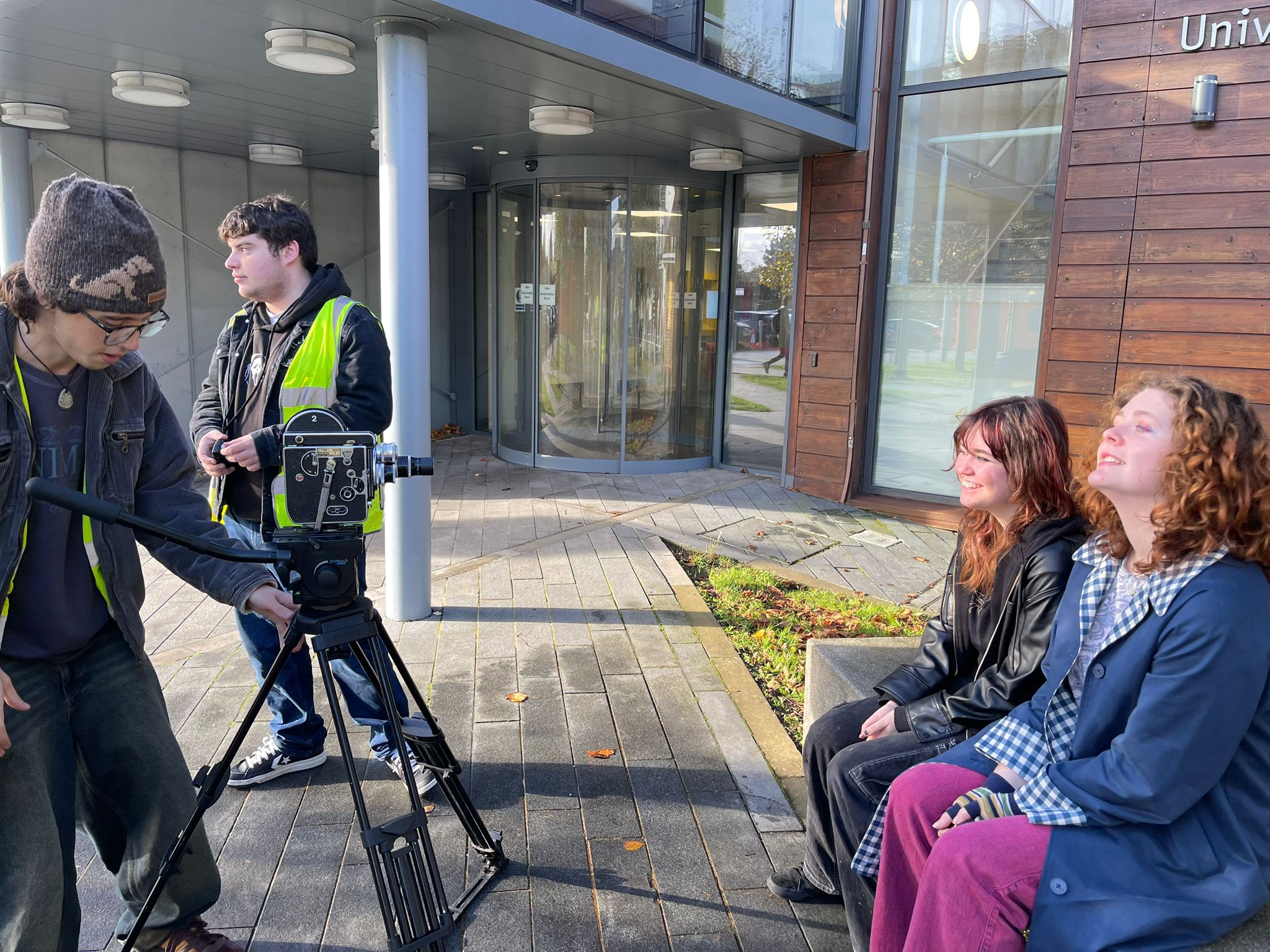 Four students outside University House on campus, two stood up next to a large film camera on a tripod, two including Rachel sat down, smiling with dark leather and colourful clothing.