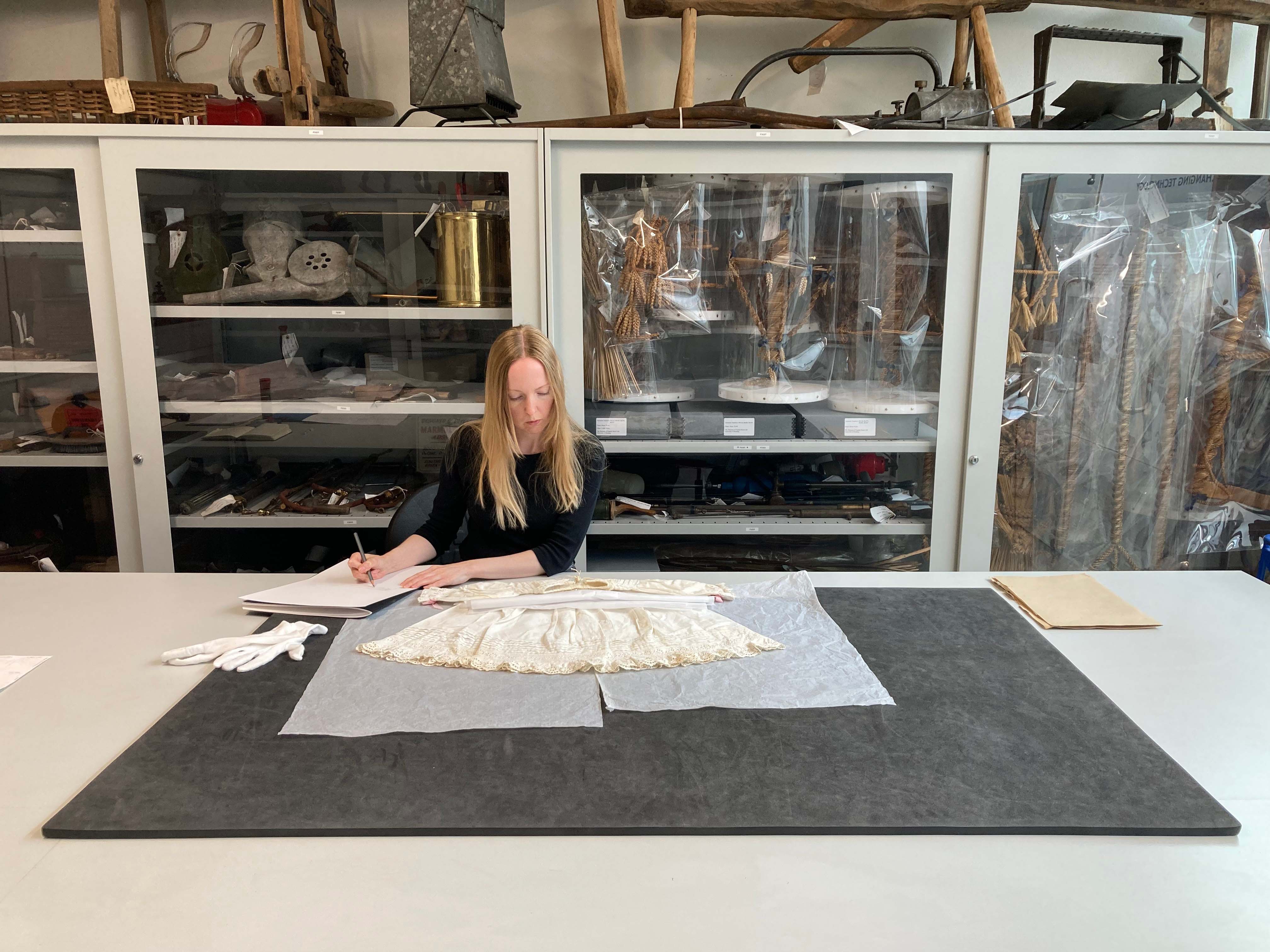 Rachel Jackson working in a studio, drawing on paper with a cream-coloured dress, large cabinets behind her.