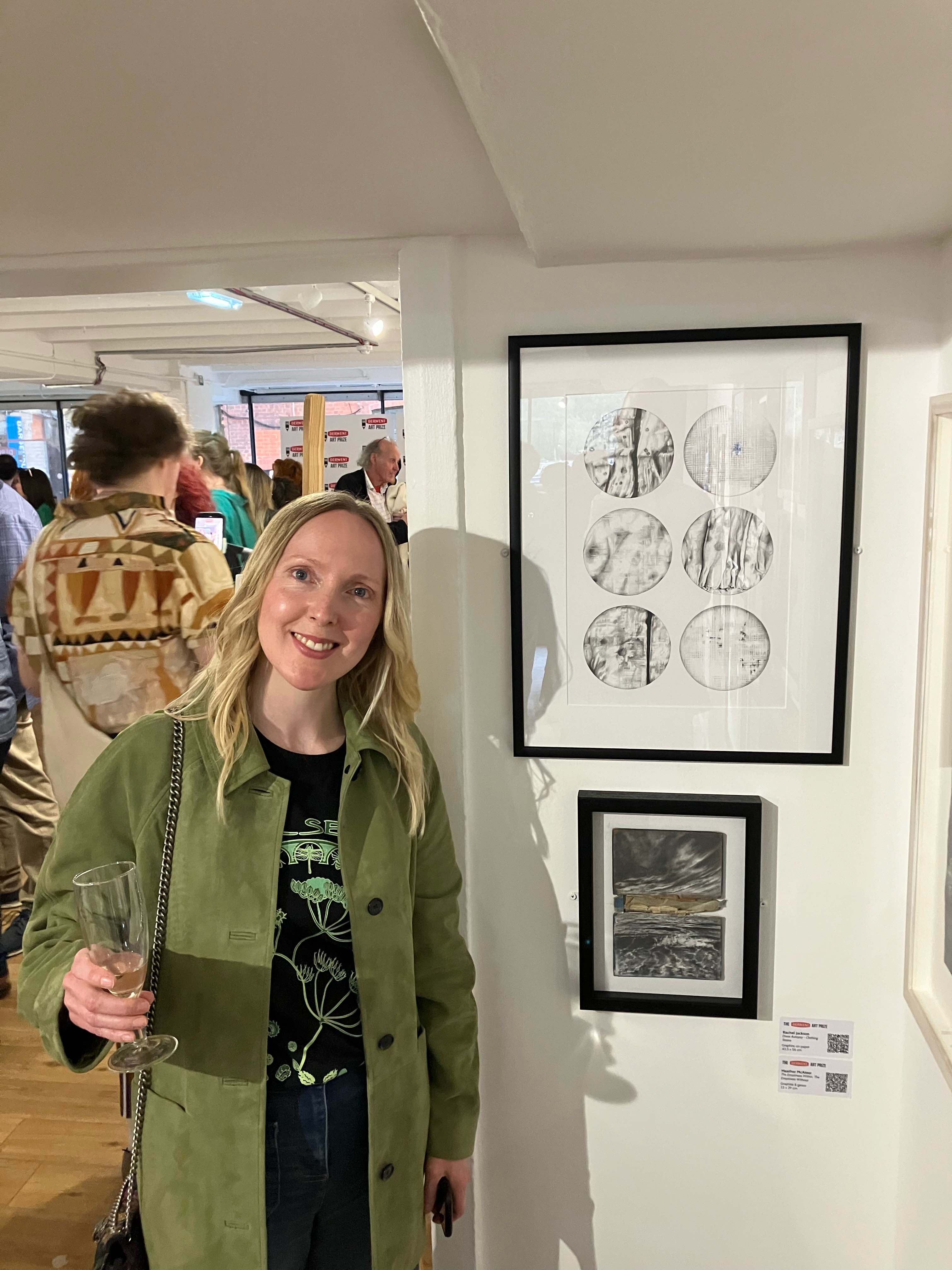 Rachel stood by her pencil-drawn work, with blonde hair wearing a green coat and holding a glass of champagne.
