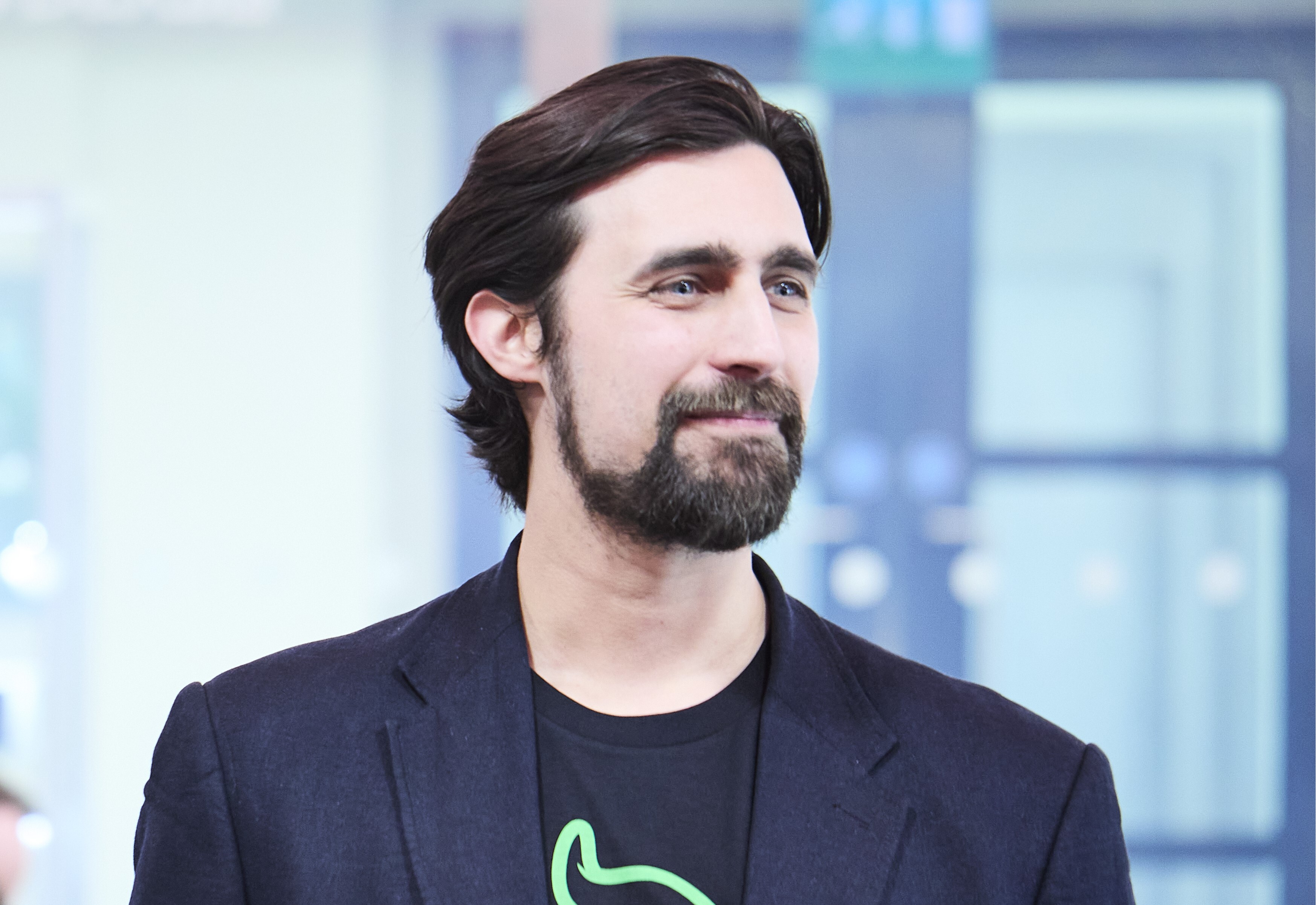 Man with dark hair and beard wearing black blazer over black t-shirt, looking to the side against blurred urban background.