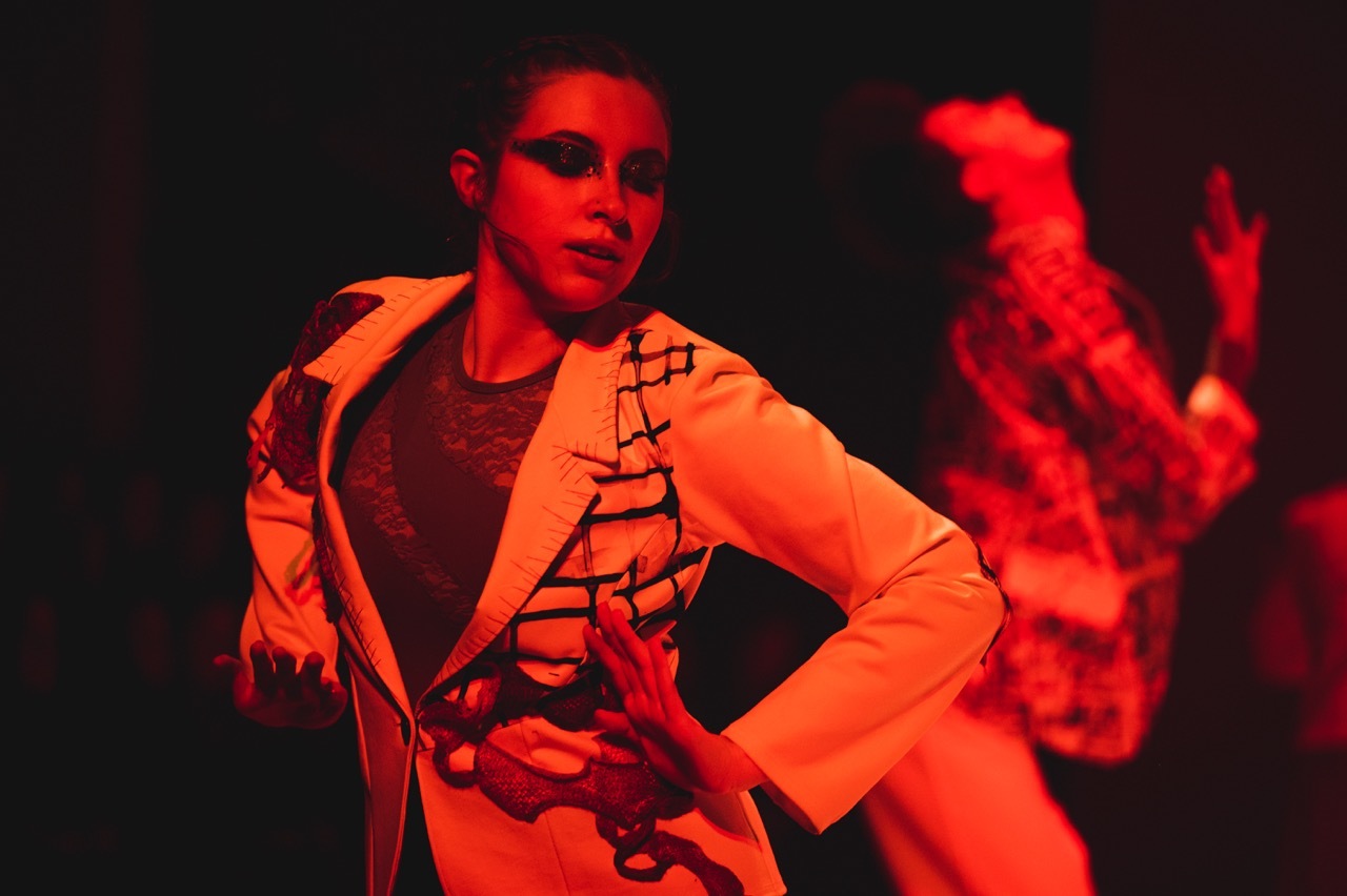 Woman with black graphic eye makeup, in a textured blazer and lace leotard from the waist up. She is on stage, lit in red and pressing her elbows sideways.