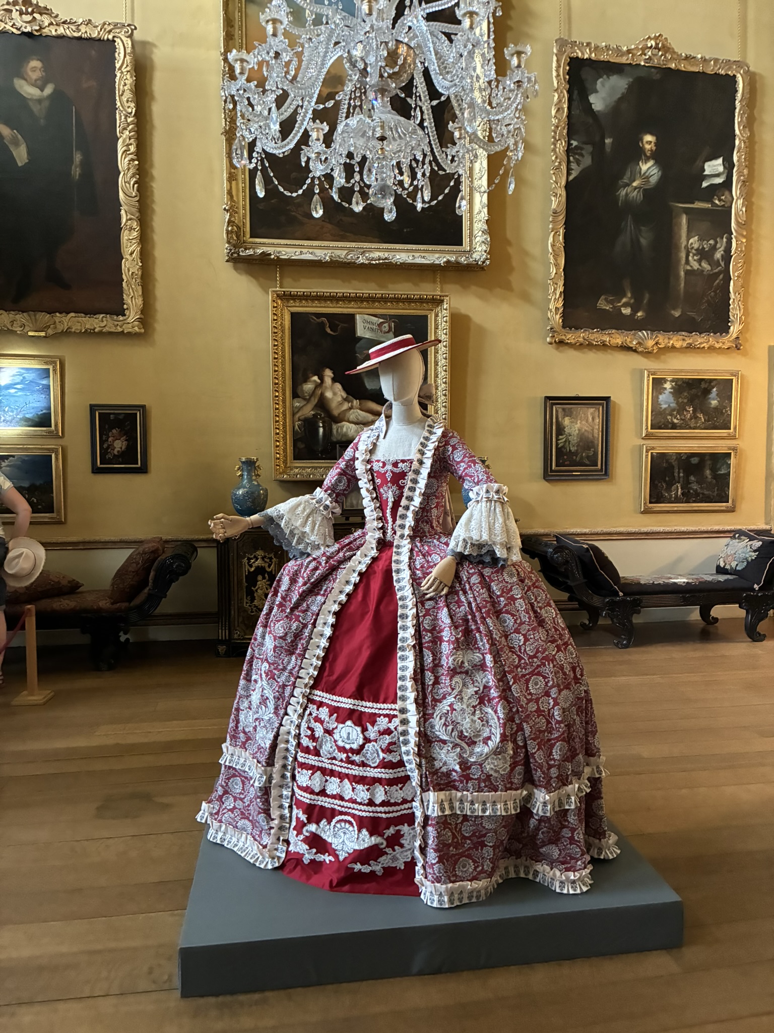 A mannequin displays an ornate red and pink historical gown on a dark plinth, with framed paintings and a crystal chandelier in a museum gallery behind it.