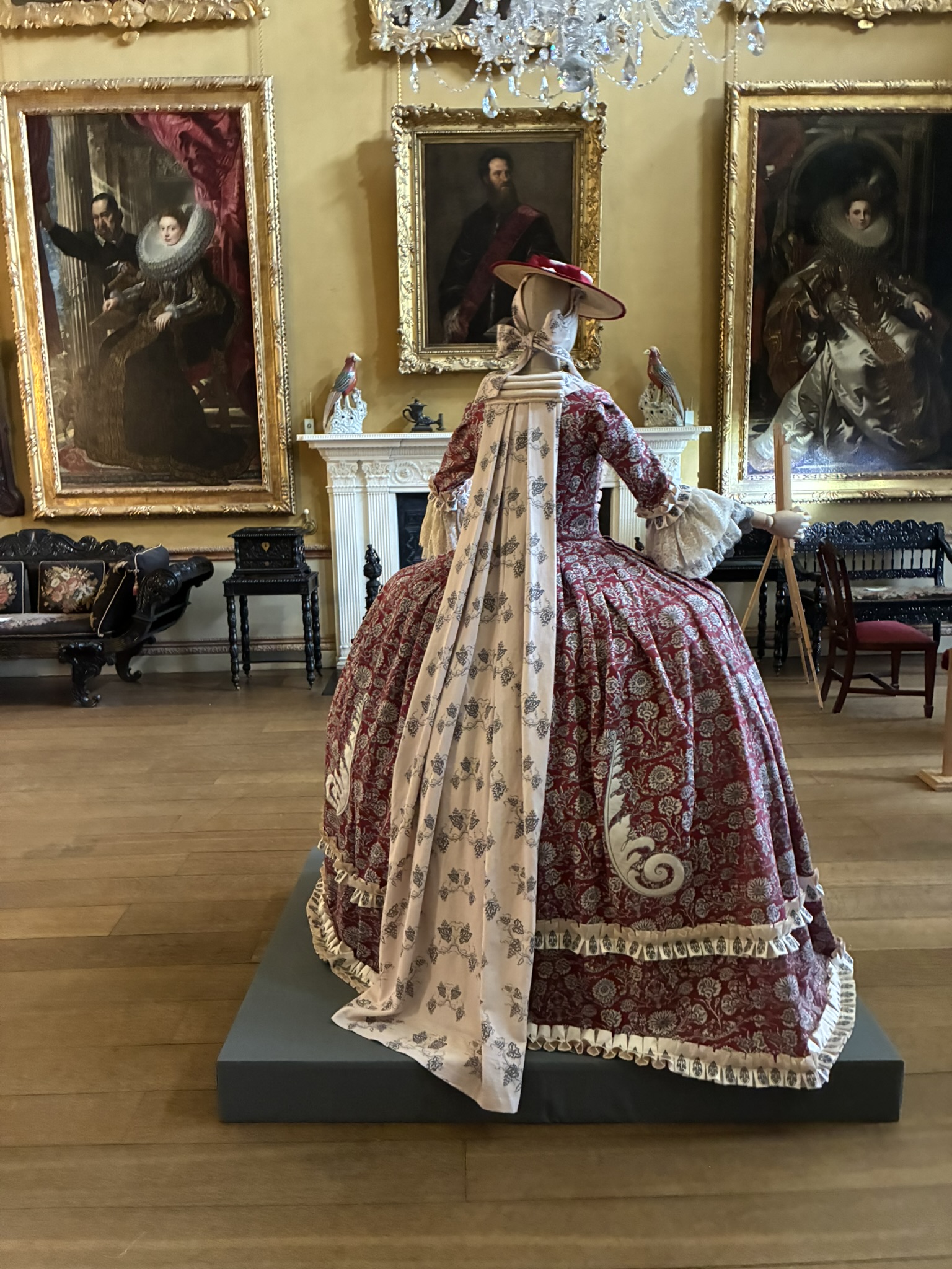 A historical dress with a full patterned skirt and long cream train is displayed on a black plinth in a gallery room with large framed portraits and a white fireplace behind it.