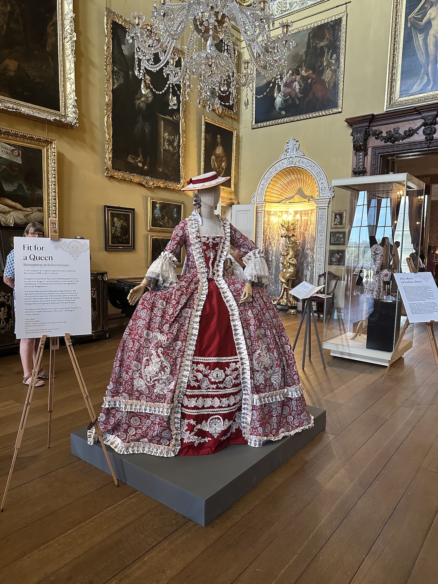 An elaborate red and cream historical dress is displayed on a mannequin in a museum gallery, with framed paintings, a chandelier and information boards in the background.