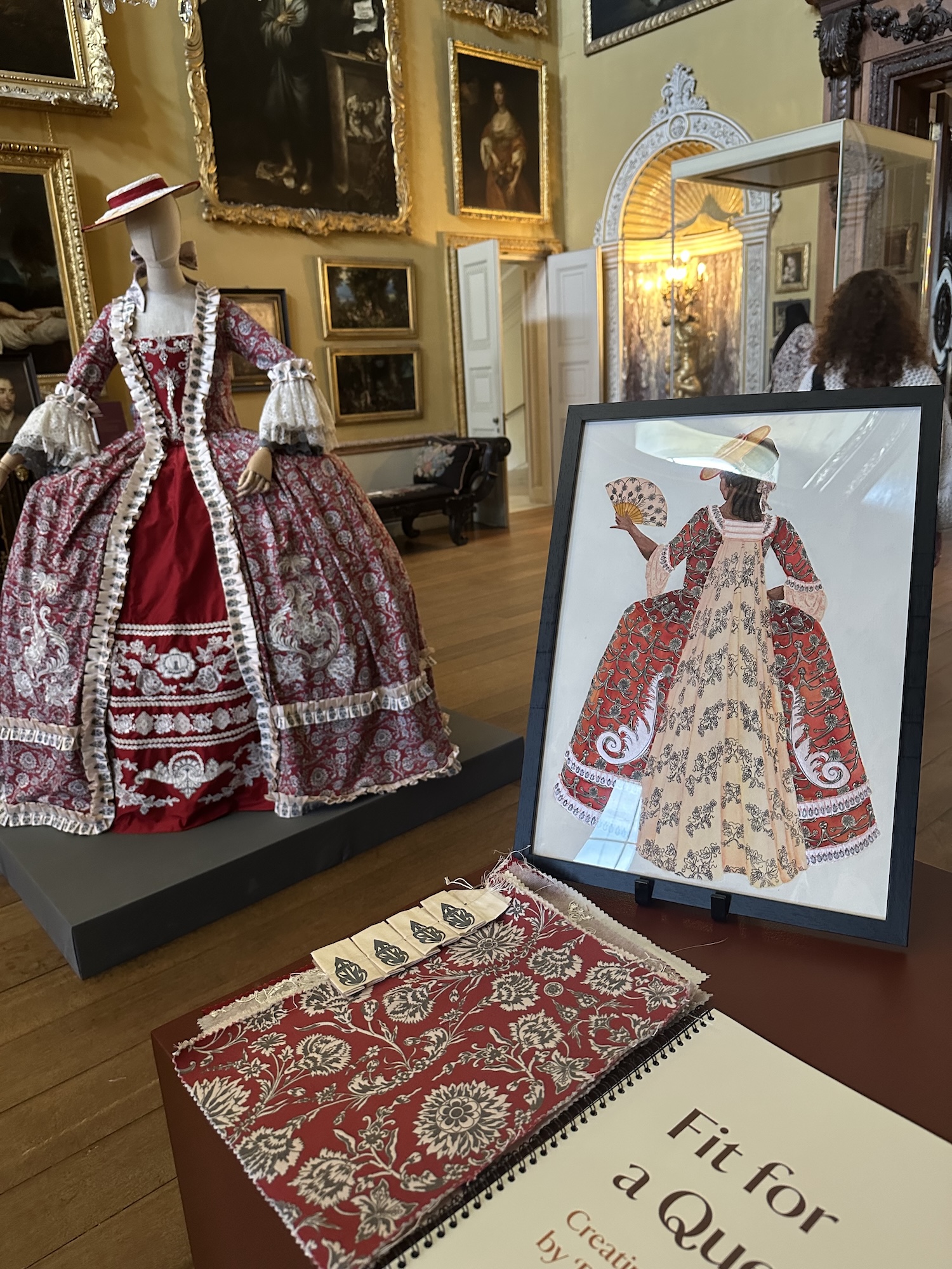 A mannequin dressed in an ornate red and white dress with matching hat in a grand room with gold-framed portraits on the wall. A book with fabric samples and a framed illustration of the ornate dress are on a table in the foreground.