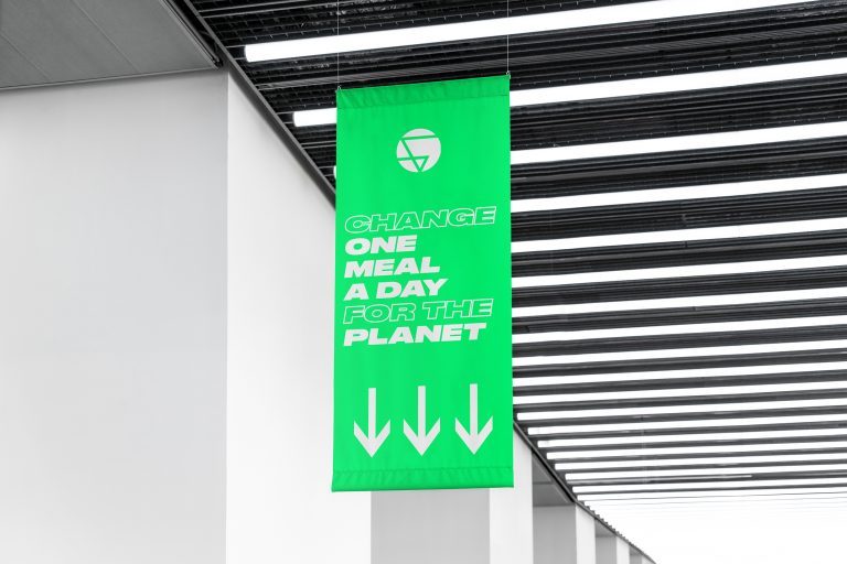 A green banner hanging from a ceiling with the text 'Change one meal a day for the planet' and three downward arrows underneath.